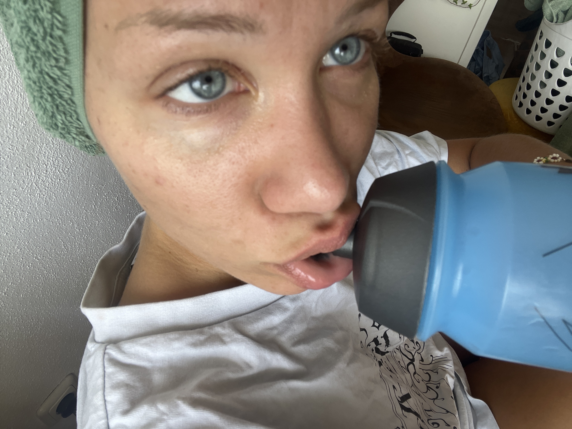 This media file captures a close-up shot of a person's face as they drink from a blue water bottle. The subject is wearing a green headband and a white t-shirt. Their eyes are open and focused, with a slight glimmer that could suggest bright lighting. The background is slightly out of focus, showing hints of a domestic setting with what appears to be furniture and a white laundry basket. The overall lighting suggests it could be daytime, possibly indoors, given the ambient light. There is no visible text in the image. The act of drinking suggests the person is rehydrating, possibly after physical activity or simply during their daily routine. The close framing emphasizes the act of consumption and the texture of the water bottle and the subject's skin.