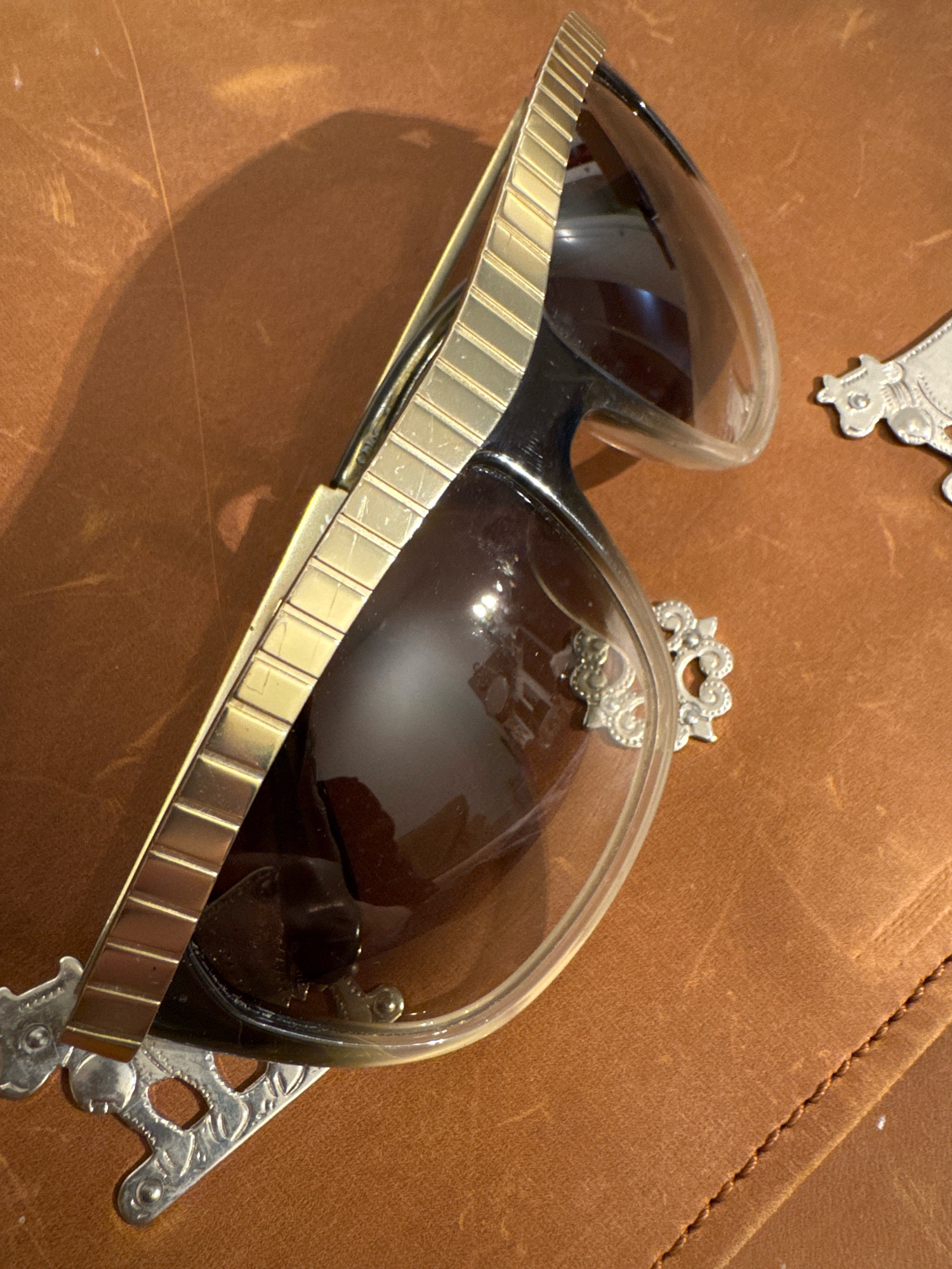 The image shows a close-up, overhead view of a pair of ornate sunglasses resting on a brown, textured surface, possibly leather. The sunglasses have a clear frame with tinted lenses and feature decorative silver accents on the sides. A gold-colored detail runs along the top of the frame, composed of small rectangular segments. To the right of the sunglasses, a small, silver decorative charm shaped like a cow is visible. The lighting casts a shadow of the sunglasses onto the surface. The image does not provide any information about the city of Tomilino, Russia, nor does it depict any people, actions, or interactions.