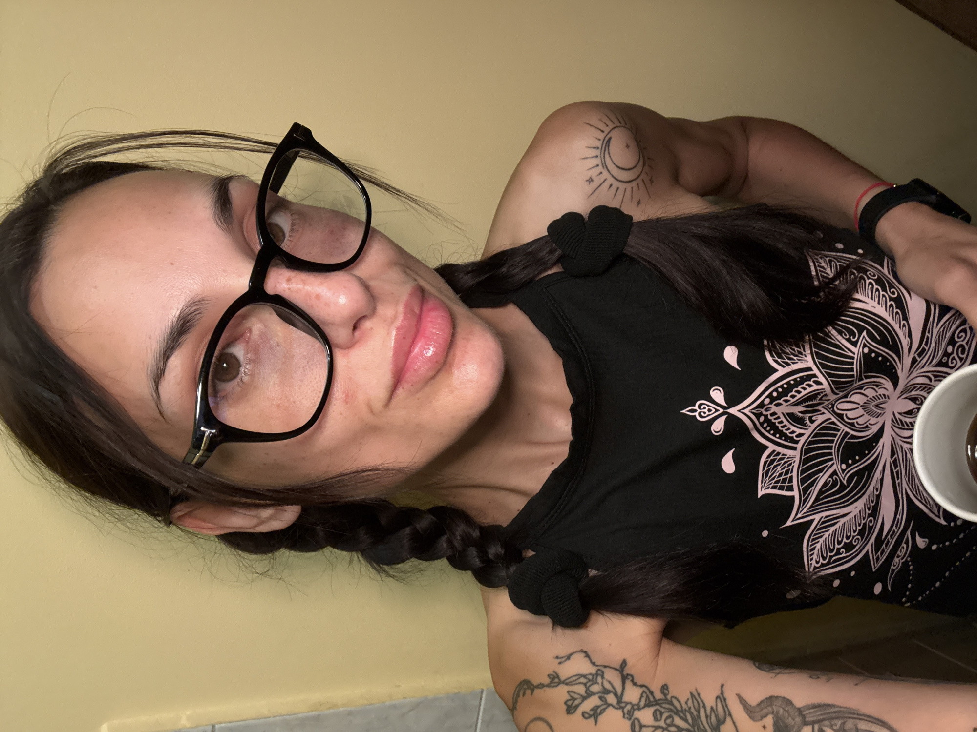 A fair-skinned woman with dark hair, styled in pigtails secured with black hair ties, is facing the camera and wearing black-framed glasses. She is wearing a black tank top with a pink floral design. A tattoo of a sun and moon is visible on her left shoulder, and another tattoo is visible on her left forearm. She is holding a white cup. The background is a pale yellow wall. The location is unknown, and there is no indication the image was taken in Pleven, Bulgaria.