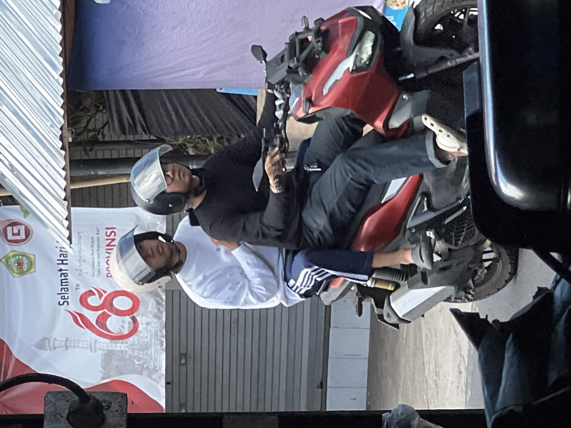 Two individuals are riding a red scooter in Payangan, Indonesia. The person in the front is wearing a white long-sleeved shirt with blue and white striped pants, and a white helmet. The person in the back is wearing a black long-sleeved shirt and dark pants, and a black helmet. The scooter has a prominent silver exhaust pipe. In the background, a banner with Indonesian text and a graphic of a tower is visible, along with a corrugated metal roof and a blue tarp. The setting appears to be outdoors, possibly along a street or in a marketplace.
