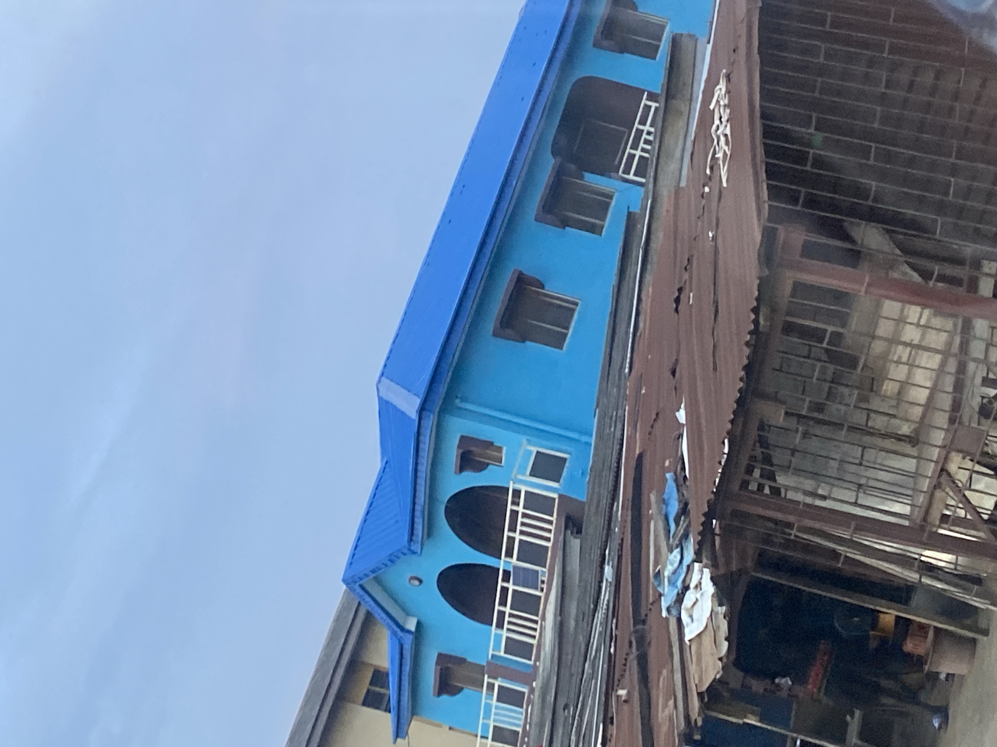 The image captures a scene in Ikorodu, Nigeria, featuring a vibrant blue building with a matching blue corrugated metal roof. The building has arched openings and a white railing on a balcony. In front of it, there is a lower structure with a rusty corrugated metal roof, appearing to be a stall or shop. This lower structure has metal bars and some items stored inside and on top of it, including what looks like blue and white tarpaulin and fabric. The sky above is overcast and a pale blue. The foreground is dominated by the rusted roof of the stall, obscuring the ground level of the blue building.