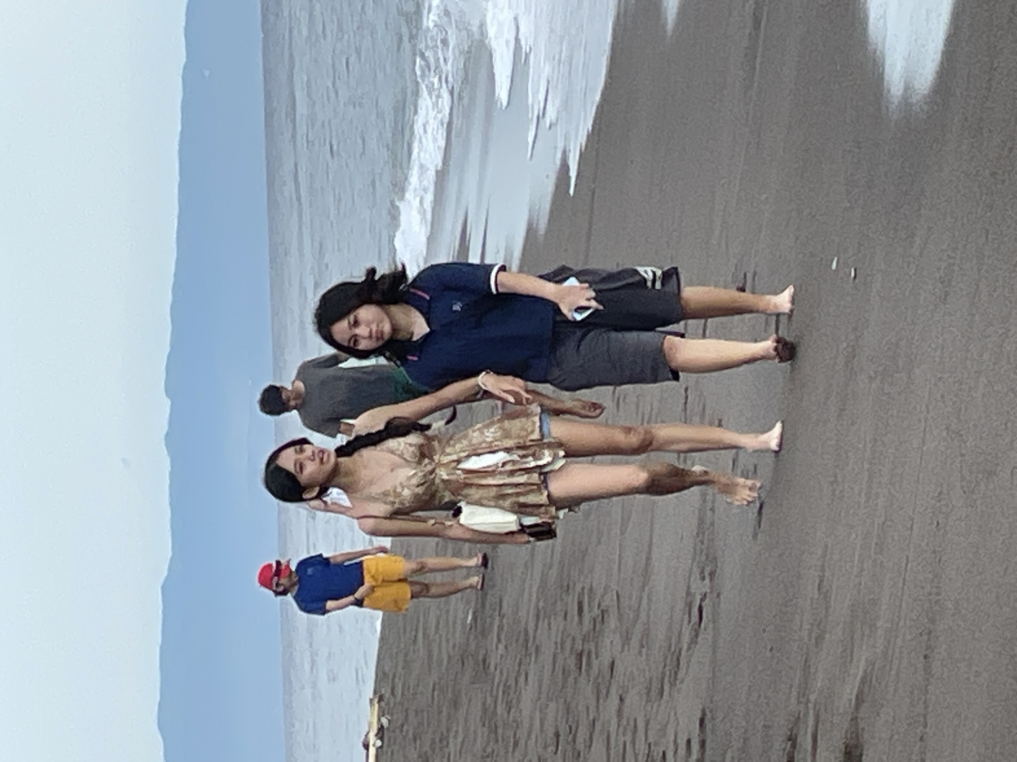 The image depicts a beach scene in Dauin, Philippines. In the foreground, two young females are walking along the dark sand, with the ocean and waves on their right. The female on the left wears a patterned, sleeveless dress and is carrying a white bag and a phone. The female on the right is wearing a dark blue polo shirt with white trim and dark shorts, and she is also holding a phone. In the middle ground, a male wearing a gray t-shirt and a person in a blue t-shirt and yellow shorts are walking further down the beach. In the background, a hazy mountain range is visible against a light sky.
