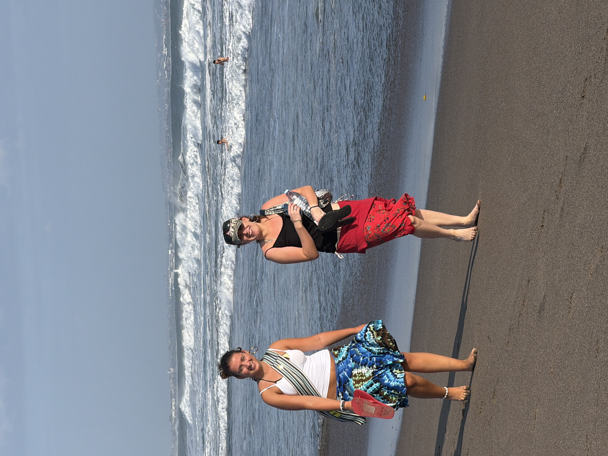 In North Kuta, Indonesia, two women walk on a dark sand beach next to the ocean. The woman on the left wears a white tank top, a blue and black patterned skirt, and holds red flip-flops. The woman on the right wears a black tank top, a red patterned skirt, sunglasses, and a cap. She carries a patterned bag and a water bottle. The ocean has moderate waves. The sky is overcast.