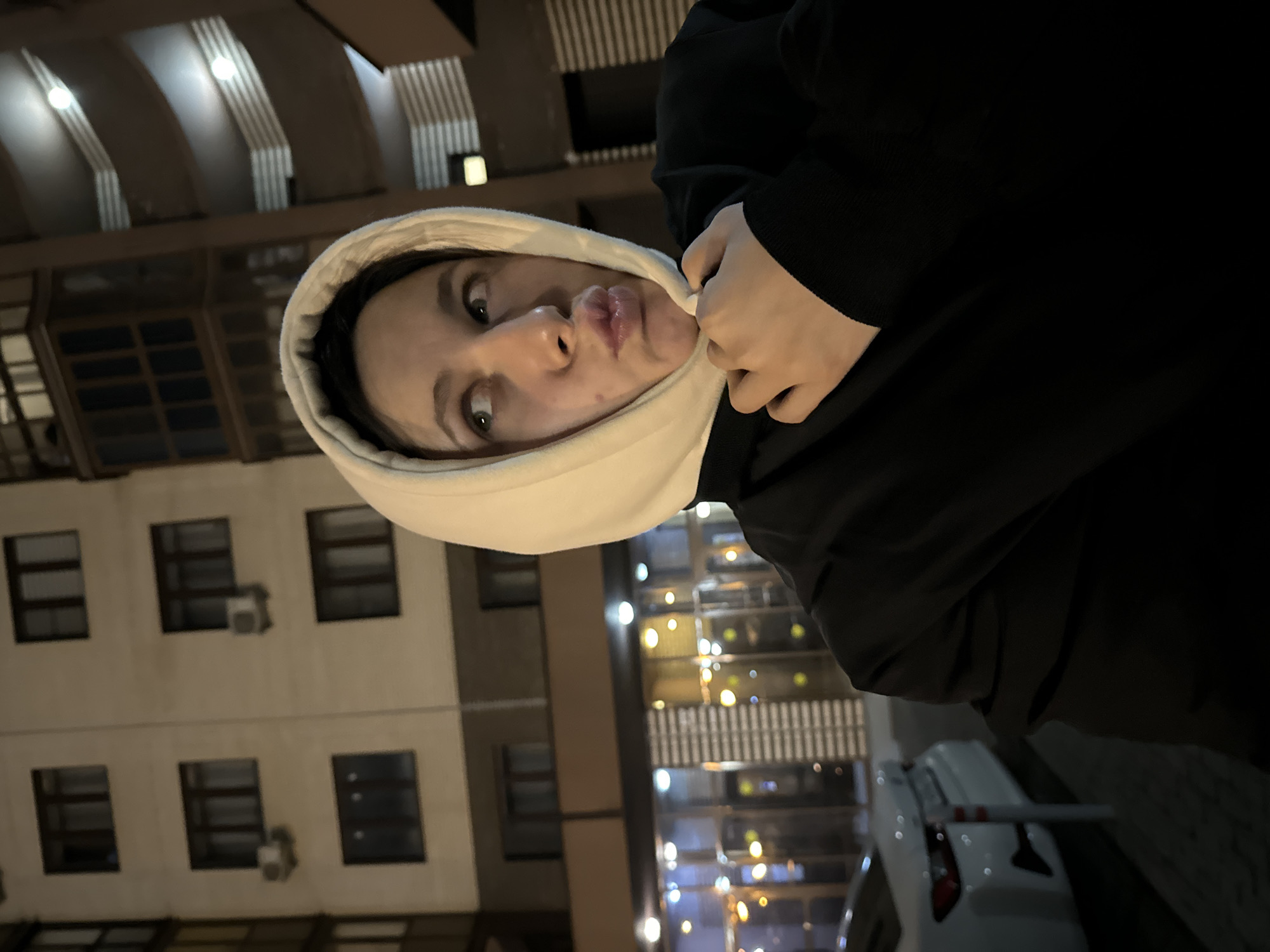 A close-up, chest-up portrait depicts an individual, presumably female, posing at night in Odintsovo, Russia. The person is wearing a light-colored hood pulled over the head and a dark garment, with the right hand holding the hood near the chin. The subject's face is turned slightly to the right, eyes looking upwards and to the right, and lips are pursed in a pouting expression. The background features an urban environment with artificial illumination. To the left stands a multi-story building with a light-toned facade, multiple dark-framed windows, and two external air conditioning units. Its ground floor exhibits brightly lit commercial storefronts with vertical metallic paneling. To the right, another building shows curved, tiered balconies or walkways illuminated by circular light fixtures. A portion of a white car and a red and white striped bollard are visible on a paved surface in the lower left of the frame.