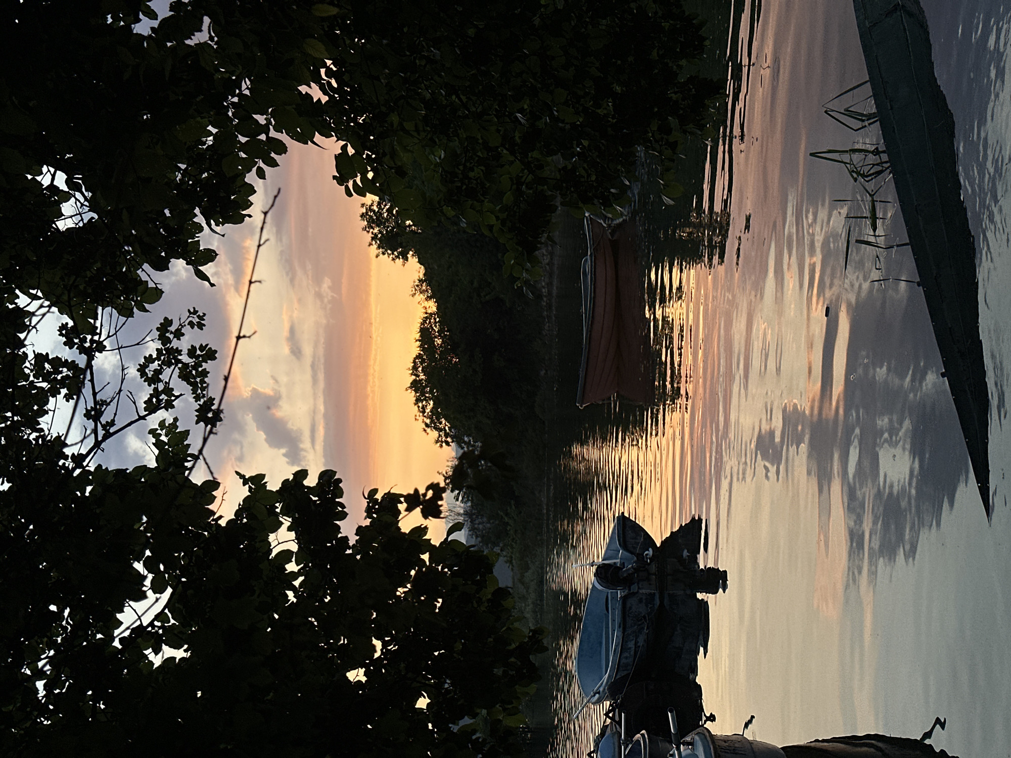 This media file captures a tranquil lakeside or riverside scene at either sunset or sunrise, likely in Glavatartsi, Bulgaria. The sky is a prominent feature, displaying a gradient of warm oranges and yellows near the horizon, fading into soft purples and blues towards the top, with scattered clouds illuminated by the low sun.
The view is framed by dense, dark foliage, primarily trees and bushes, which are silhouetted against the bright sky on the left and top-left of the image. Their dark forms create a natural opening to the water and the distant shore.
The calm water acts as a mirror, reflecting the vibrant colors of the sky and the dark shapes of the surrounding trees, creating elongated, wavy patterns of light and shadow. A small, light-colored boat is partially visible on the water, seemingly moored or drifting gently, its form subtly defined by the reflections.
On the right side of the image, what appears to be a dark bank or dock extends into the foreground. Near this edge, reeds or tall grasses emerge from the water. Another dark, indistinct object, possibly part of a boat or watercraft with some mechanical elements, is visible in the extreme foreground on the right, heavily shadowed.
The overall atmosphere is one of serene peacefulness, characteristic of the golden hour. There is no activity taking place, and no people are visible. No text is visible in the image.