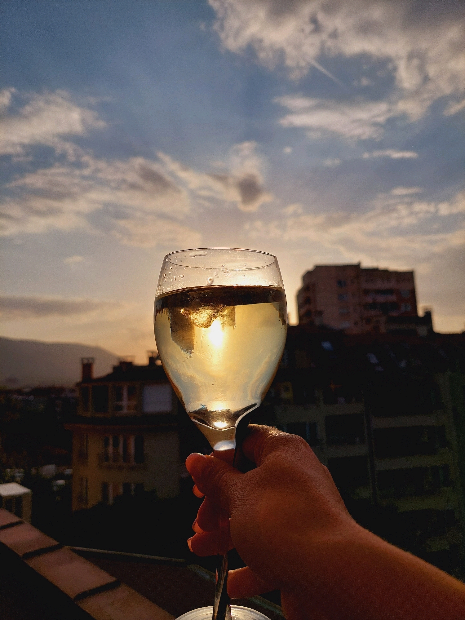 A hand holds a wine glass filled with a light-colored liquid, likely white wine, against a backdrop of a city skyline during sunset. The sun's rays are visible, reflecting off the surface of the liquid and casting a warm glow. In the background, several buildings of various heights and architectural styles are silhouetted against the sky, with some lights beginning to appear in the windows. A mountain range is faintly visible in the distance. The sky is filled with clouds, tinged with the orange and yellow hues of sunset. The scene appears to be from a balcony or rooftop in an urban setting, possibly Sofia, Bulgaria, given the architectural style of the buildings and the mountainous horizon often seen from the city.