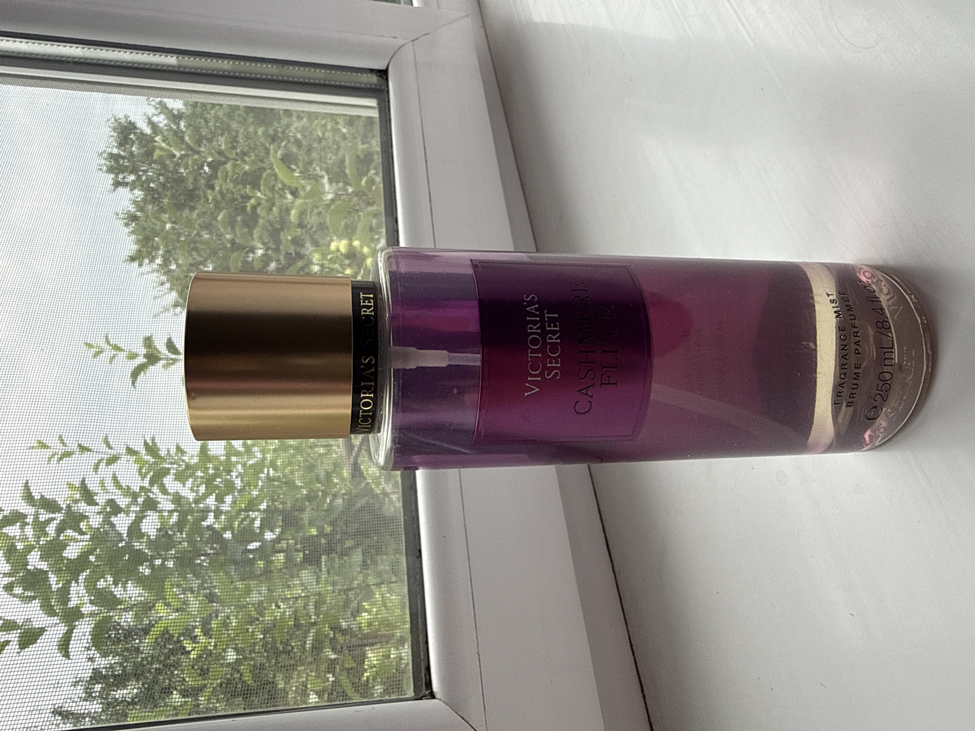 A Victoria's Secret body mist bottle, labeled "Cashmere Fleur," sits on a white windowsill against a backdrop of green trees seen through a window screen. The bottle is made of clear purple glass with a gold cap. The label is purple with white text. The scene is indoors, looking out a window, and there are no people present. The location is not specified as Tekeli, Kazakhstan.
