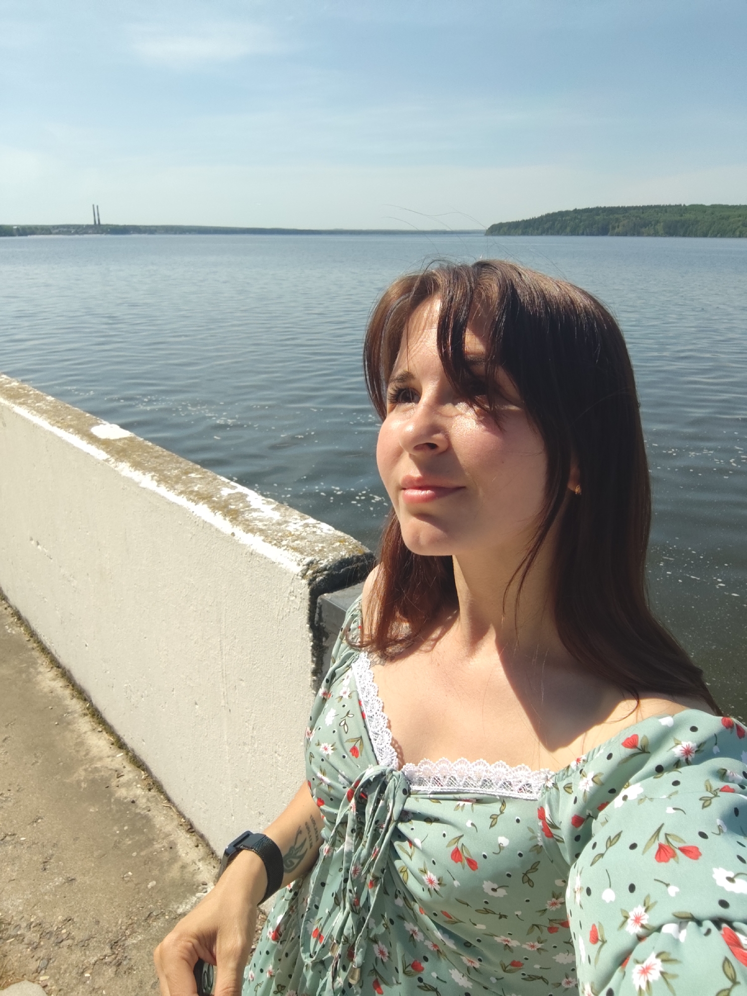 A fair-skinned woman with brown hair poses for a selfie on a sunny day. She stands next to a low, white concrete wall, with a large body of water behind her. In the distance, a shoreline is visible, with a pair of tall, slender structures emerging from it. The woman wears a light green floral dress with white lace trim around the neckline. A dark-colored wristwatch is on her left wrist. The location is likely the city of Chaikovsky, Russia, on the Votkinsk Reservoir.