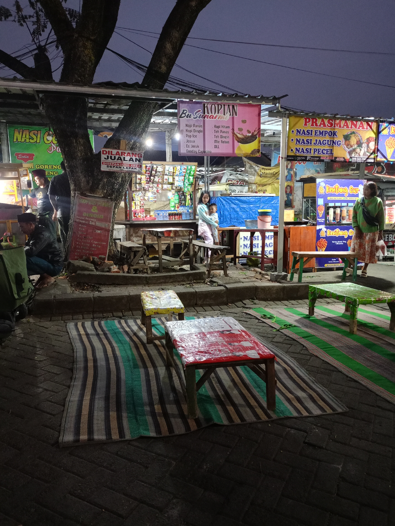 The image depicts an outdoor food stall scene at dusk or night in Donowarih, Indonesia. Several food stalls are set up under a canopy supported by a large tree. Banners advertise various food and drink options, including "NASI GORENG" (fried rice), "KOPIAN" (coffee), and "PRASMANAN" (buffet-style dishes like nasi empok, nasi jagung, and nasi pecel). There are also signs for "Kentang Curi" (stolen potatoes) and drinks.
In the foreground, two low tables with red and white coverings sit on striped mats laid out on dark brick paving. Several low stools are also visible. In the middle ground, there are more wooden tables and chairs. People are present at the stalls; a man is squatting by a stall on the left, and a woman with a child is standing at a stall in the center. Another woman is standing near a stall on the right. The environment appears to be a marketplace or roadside area with an ambient, dimly lit atmosphere. Electrical wires are visible crisscrossing overhead, suggesting an urban or semi-urban setting.