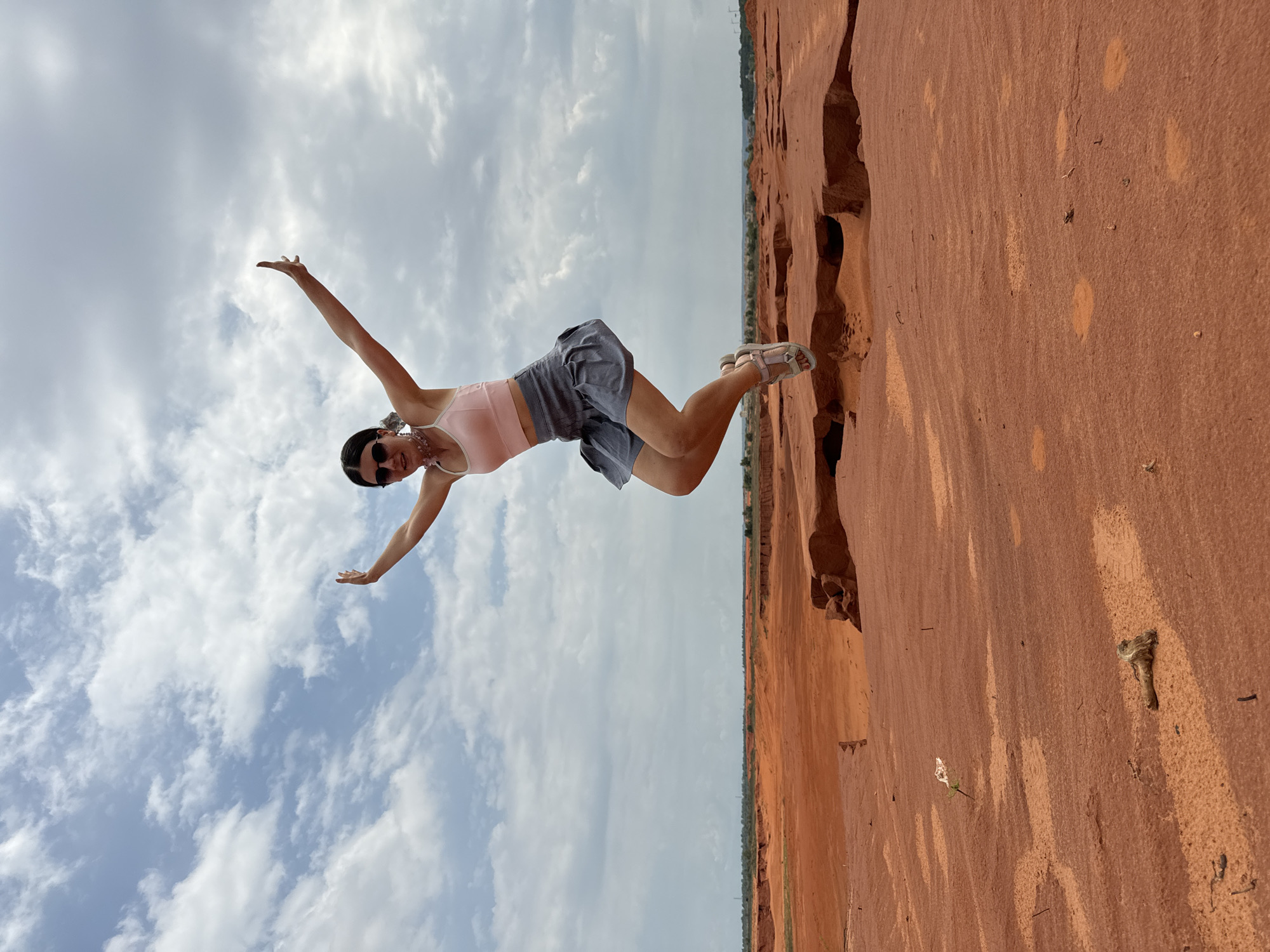 A young woman with dark hair, wearing sunglasses, a light pink tank top, and a gray skirt, is captured mid-air in a jump. She is wearing sandals and a delicate necklace. Her arms are outstretched to the sides, and her legs are bent as if in motion. The woman is positioned in the foreground of a vast, arid landscape. The ground is a vibrant reddish-orange hue, with textured patches and what appear to be natural erosion patterns forming crevices and layered rock formations. In the distance, a flat, brown horizon stretches across the image, meeting a pale blue sky filled with scattered white clouds. The scene suggests a remote, desert-like environment, consistent with the sand dunes and unique geological formations found in Mui Ne, Vietnam. There are no other individuals visible in the frame.