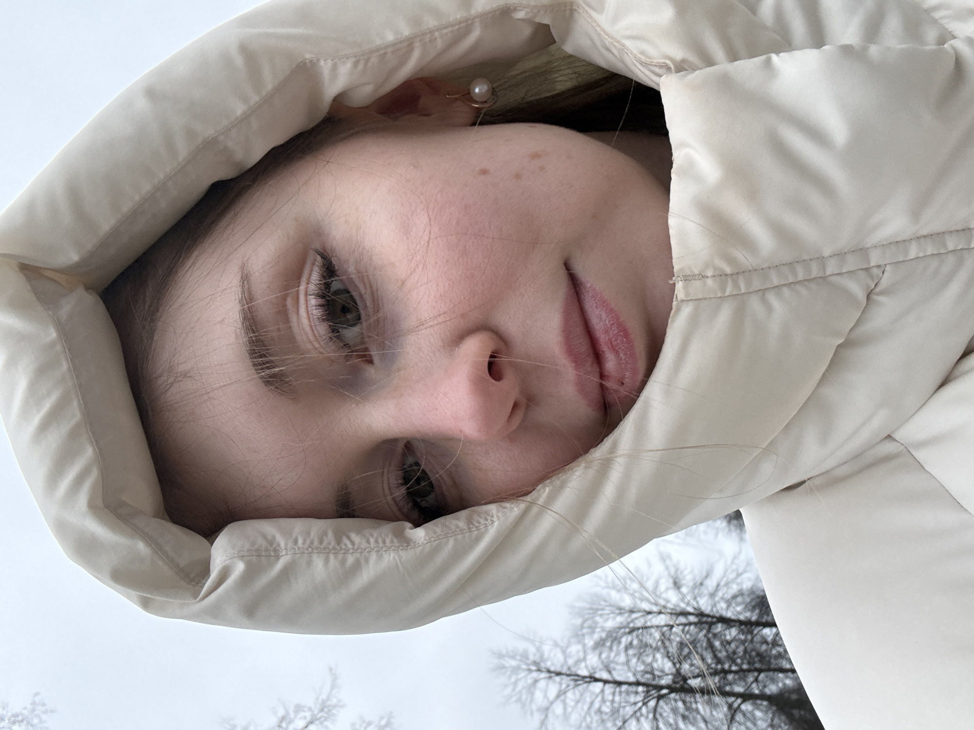 A close-up, slightly angled shot depicts a young woman's face framed by a light-colored, possibly white, puffy jacket hood. The woman has fair skin with freckles and light-colored hair. She wears a pearl earring on her left ear. Her expression is relaxed with a slight smile. The background, visible beneath the hood, features a cloudy sky and bare tree branches, suggesting a winter or late autumn scene. The location is unidentified.