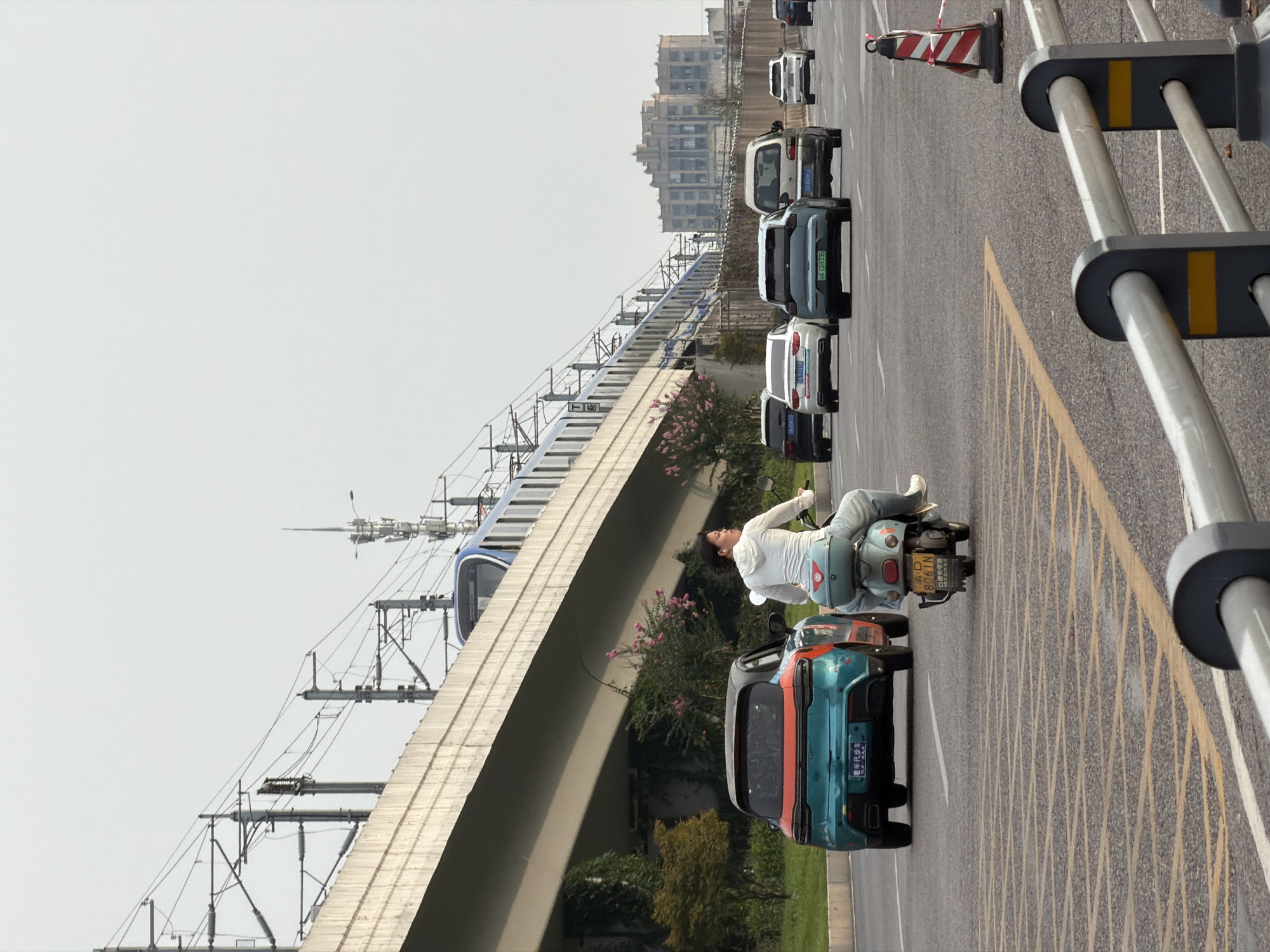 An outdoor, eye-level, medium shot shows a street scene in the city of Jiangjin, China, with a bright, overcast sky. In the foreground, a light blue scooter with a rider wearing a white hooded sweatshirt and blue jeans is traveling in the right lane of a multi-lane road. A teal and orange car is in the left lane, moving away from the viewer. In the background, an elevated train track runs parallel to the road, with a blue and white train visible on the tracks. Buildings and trees line the side of the road, and traffic cones are placed along the roadway.
