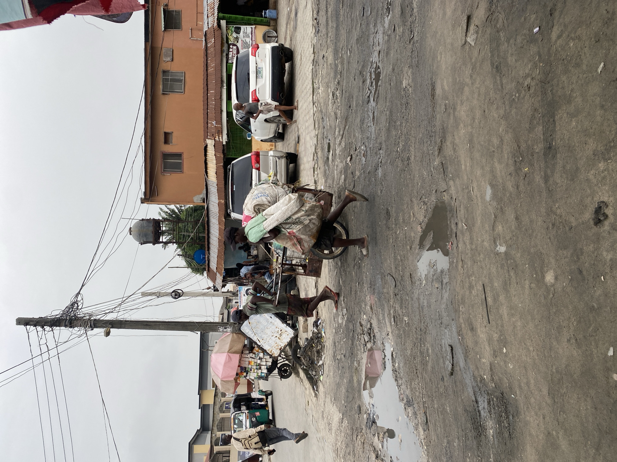 The scene captures a street in Ado, Nigeria, characterized by a worn, cracked asphalt road with puddles. A man is seen pushing a laden cart with a large sack of goods balanced on top. Another man is pushing a cart laden with goods covered by a pink umbrella. A man stands next to two parked cars near a building with a corrugated metal roof. Power lines run across the frame supported by a concrete pole. In the background is a building with multiple windows. The sky is overcast.