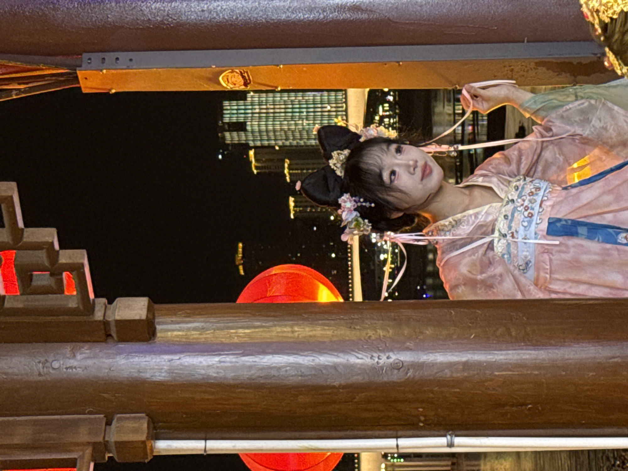 In the Yuzhong District of China, a young East Asian woman, adorned in traditional Hanfu attire, is positioned behind a wooden pillar on a waterfront at night. She has styled her hair with black bows and floral accessories. The backdrop reveals a cityscape with illuminated skyscrapers and a bridge spanning a body of water, suggesting a prominent urban setting. Red lanterns are visible, adding to the festive ambiance. The woman is looking to her right, with her right hand lightly touching a ribbon attached to her garment. The overall scene captures a blend of traditional cultural elements within a modern city environment.