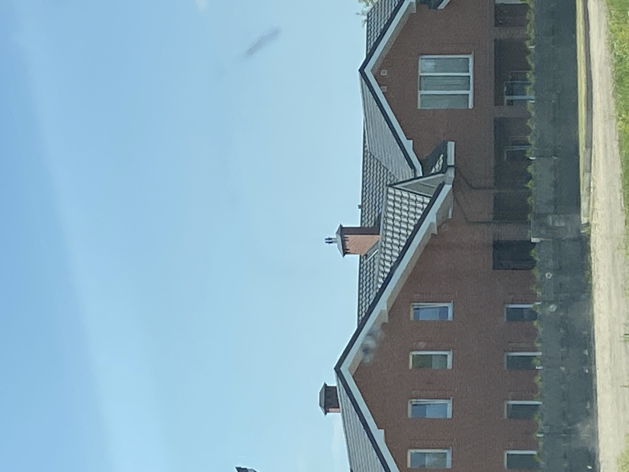 The image shows the exterior of two connected, multi-story red brick houses under a clear blue sky. Both houses have dark gray tiled roofs with white trim. The left house has at least five windows visible on its upper floor and three on its lower floor, with a dark doorway on the far right. The right house features a large, multi-paned window on its upper floor and several dark openings on its lower level. Chimneys are visible on the roofs of both structures. A low, dark green fence with visible foliage behind it runs along the front of the properties. The ground in front of the fence appears to be a dirt or gravel path, with some green grass in the foreground. The scene is likely located in Матыкальскі сельскі Савет, Belarus.