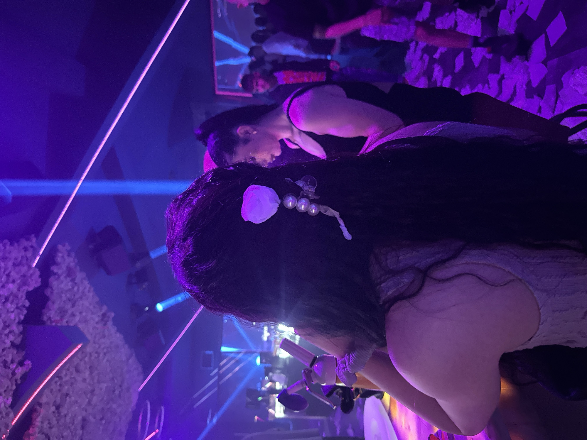 The image depicts an indoor scene within a nightclub or bar, specifically in the city of Sliven, Bulgaria, characterized by vibrant purple and blue ambient lighting. In the immediate foreground, viewed from the back, is a person with long, dark hair. Their hair is adorned on the right side with a white floral accessory and a string of three white pearls. This individual is wearing a light-colored, possibly white or grey, sleeveless top with a textured pattern and has their right arm bent, resting on an unseen surface, likely a bar counter.
To the right of the foreground person, another individual is seen in profile, facing left. This person has dark hair tied in a high ponytail and is wearing a plain black tank top. They appear to be standing near the foreground person, possibly at the same counter.
The background reveals a blurred crowd of multiple individuals, indicating a social gathering. A male figure wearing a dark jacket over a red graphic t-shirt is partially visible, looking towards the right. The floor in the mid-to-background on the right side is covered with numerous crumpled white papers, resembling confetti or discarded items. A person's lower legs and feet, wearing dark sneakers, are visible amidst this debris.
The ceiling features decorative elements, appearing to be clusters of light-colored, possibly artificial, flowers or foliage. Linear light strips emitting pink and purple light traverse the ceiling and walls, while beams of blue light cut through the smoky or hazy air, contributing to the club atmosphere. A structure in the deeper background displays illuminated blue horizontal lines. On the left side of the foreground, a white, metallic object, resembling a whipped cream dispenser or similar bar equipment, is visible.