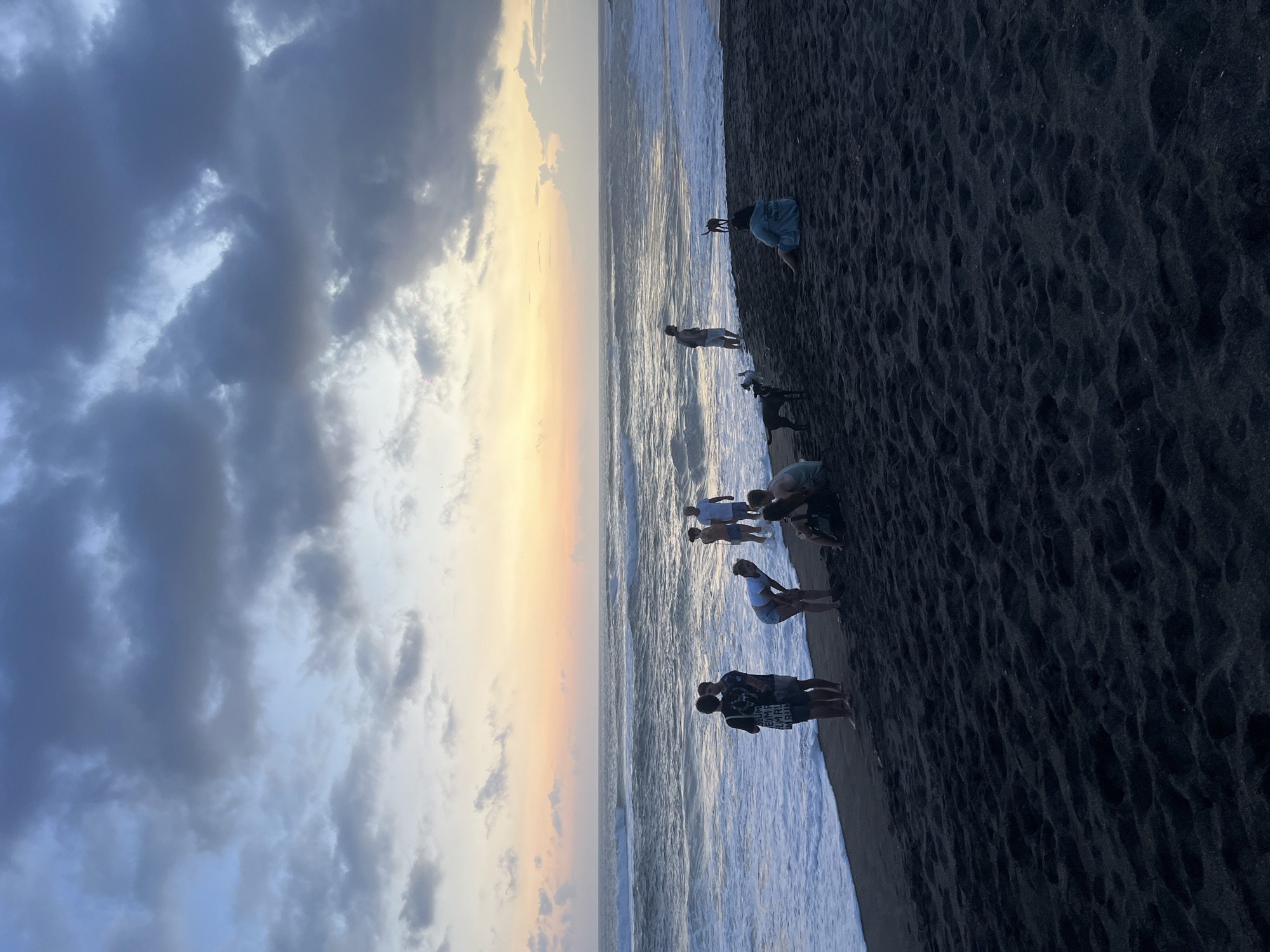 A coastal scene unfolds at dusk in Mengwi, Indonesia. The sky displays a gradient of clouds transitioning from dark grey to a light yellow-orange hue. Several people are situated on a black sand beach, some standing and others bending down near the water's edge. The ocean waves are white-capped as they approach the shore.