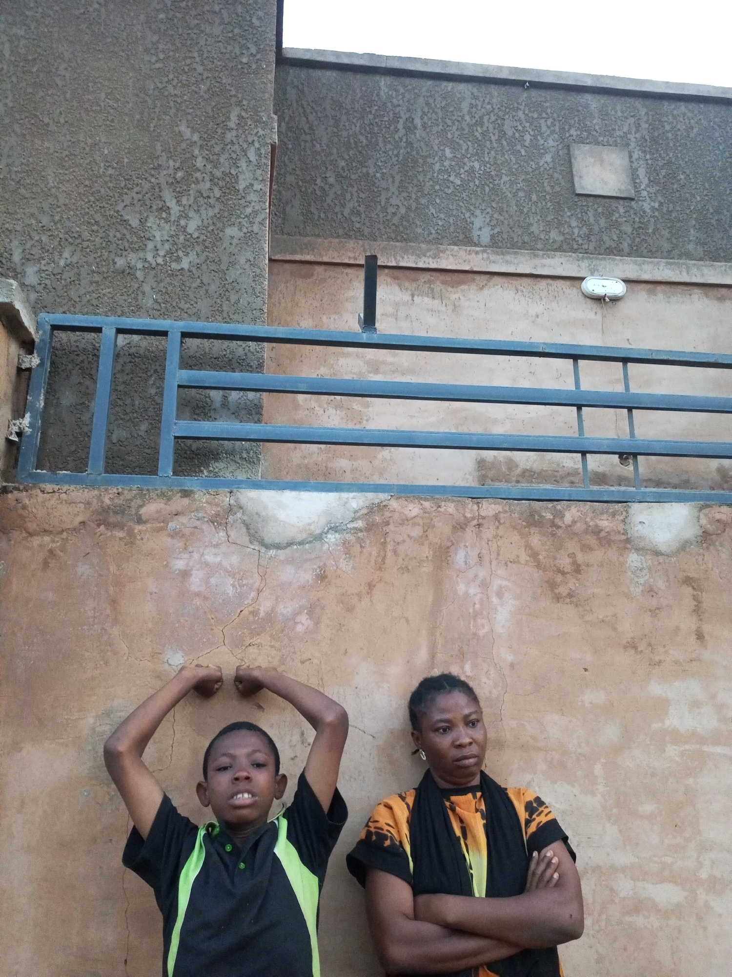 A medium shot depicts two individuals posing in front of a weathered, textured concrete wall. On the left, a young boy, appearing to be of Nigerian ethnicity, stands with his arms bent at the elbows, fists clenched and raised towards his head, flexing his biceps. He wears a black short-sleeved shirt with neon green accents on the collar and sides. His facial expression is a slight grimace. To his right, a woman, also appearing to be of Nigerian ethnicity, stands with her arms crossed. She has braided hair and wears a black scarf around her neck and a brightly colored orange and yellow patterned short-sleeved shirt. Her expression is neutral, and she gazes off to the side. The environment suggests an outdoor setting, likely in the city of Tempure, Nigeria. A dark blue metal railing runs horizontally across the middle ground of the image, positioned between the wall and the individuals. Above the railing, a gray textured upper wall is visible, with a square fixture and an oval light fixture mounted on it. The lower wall exhibits signs of age and wear, with patches of peeling paint and visible cracks. The lighting suggests it is daytime, with overcast conditions.