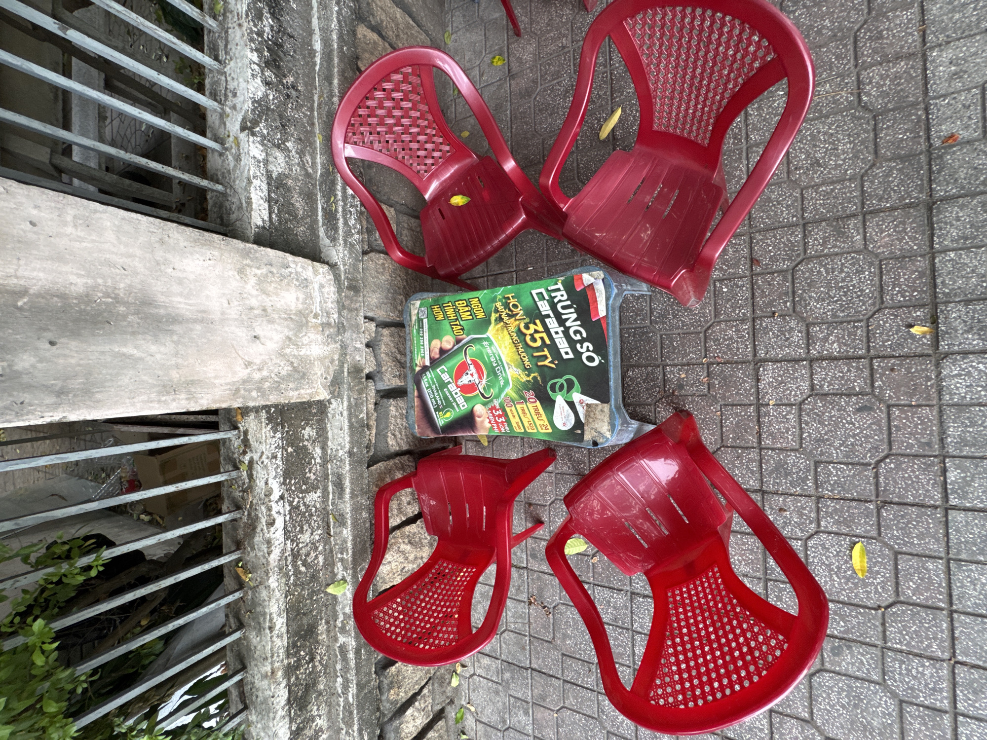 This is a ground-level, outdoor, eye-level shot of an informal seating area in Nha Trang, Vietnam, likely on a sidewalk. Four bright red plastic chairs are arranged around a low, blue plastic table on a patterned gray tiled surface. The table has a crumpled, bright green and yellow advertisement for "Carabao" energy drink, with Vietnamese text. The chairs are simple, modern designs with perforated seats and backs, and armrests. A weathered concrete wall runs along the top of the frame, behind a metal fence that encloses some foliage and what appears to be discarded cardboard boxes. Scattered yellow leaves are visible on the ground and on the chairs. The scene suggests a casual, potentially temporary, setup for outdoor refreshments.