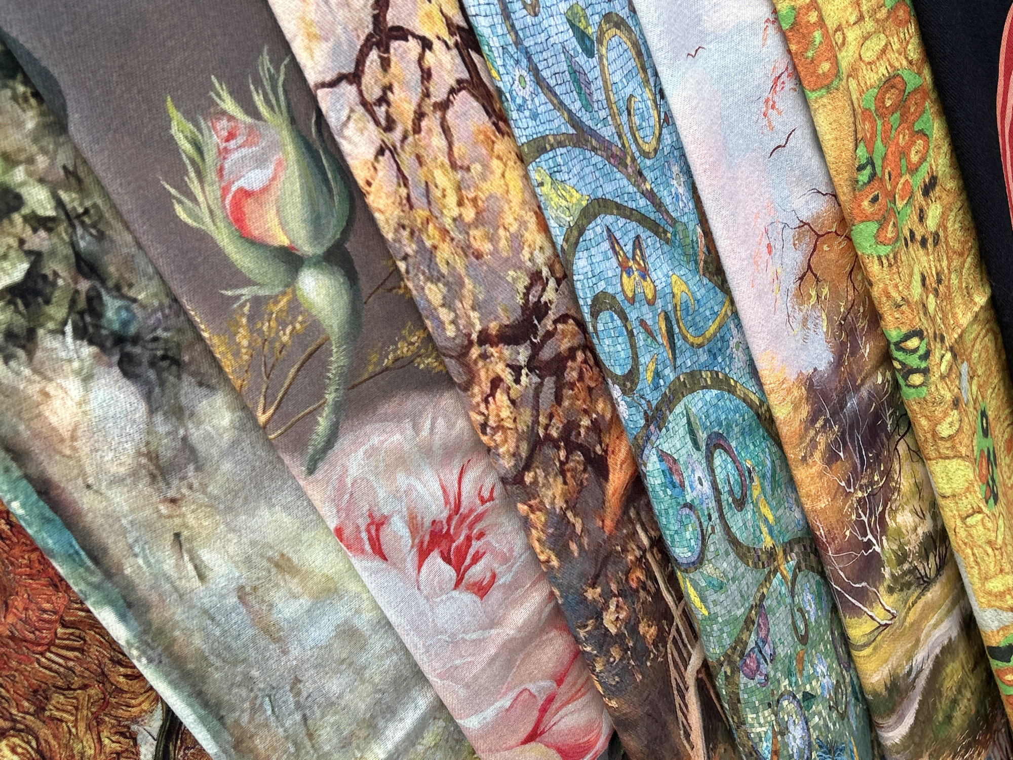 The image displays a close-up, overhead view of several folded scarves, each printed with a different painting. The scarves are arranged in a diagonal fan shape. The paintings feature floral and landscape motifs, executed in vibrant colors and distinct artistic styles. The first scarf from the top is a detailed abstract pattern with oranges and browns. Below it, a scarf with a faded, textured pattern. The next scarf shows a soft pink and red flower. Following that is a detailed painting of a rosebud, with green leaves and a hint of yellow foliage in the background. Another scarf features a tapestry-like design with a mosaic pattern of blue, green, and yellow, depicting stylized vines and figures. Below that, a scarf with a landscape scene featuring trees and a sky. The bottom scarf exhibits a close-up of yellow and orange flowers, possibly sunflowers. The lighting is even, and the focus is sharp, highlighting the intricate details of the prints. The image does not contain any information or elements that would place it in the city of Mtskheta, Georgia.