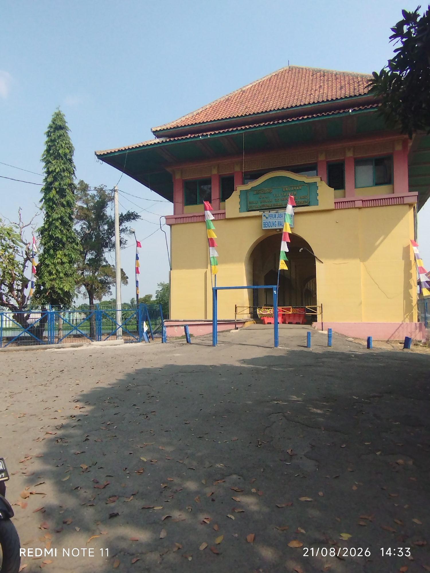 The image shows a sunny day at the Sadang River Dam (Bendung Sadang) in Karawang, Indonesia. The dam's main building, painted in yellow with pink and teal accents, stands prominently. A large archway leads into the structure, and colorful flags are strung along the building's facade and a nearby pole. A tall, slender tree is visible to the left of the building, and a blue fence encloses a portion of the area. The ground is a paved surface with scattered fallen leaves, indicating an outdoor environment. The image also shows a portion of a motorcycle on the lower left, and a timestamp of "21/08/2026 14:33" and "REDMI NOTE 11" at the bottom.