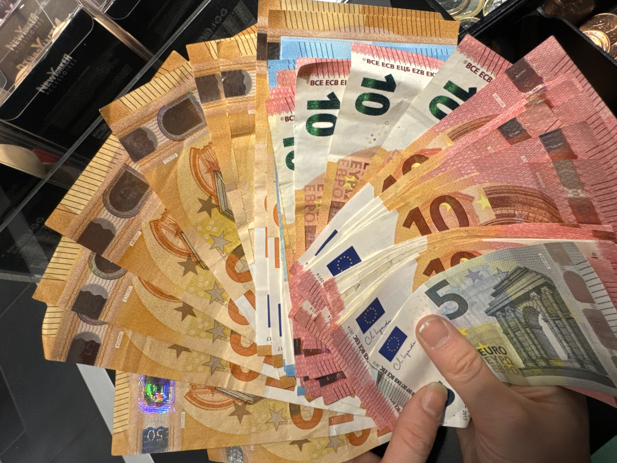 A hand holds a fan of Euro banknotes. The denominations visible include 50 Euro, 10 Euro, and 5 Euro notes. The banknotes are fanned out over a black surface, next to a plastic display case with "New Yorker ACCESSORIES" printed on it. Some coins are visible in the background.