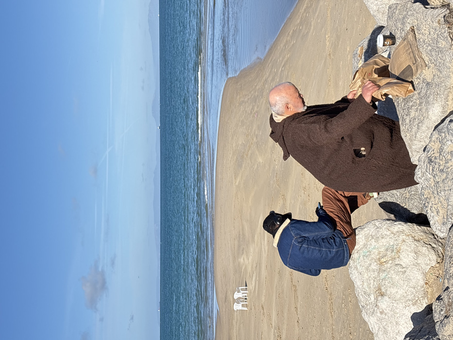 The image depicts a bright, clear day on a sandy beach in Tangier, Morocco. In the immediate foreground, a cluster of large, light-colored rocks forms the base of the scene. Two individuals are present among these rocks. On the left, a person, seen from the back, is seated on a rock. This individual wears a dark blue denim jacket with a light-colored shearling collar, rust-colored pants, and a dark bucket hat, and is looking down at a mobile phone held in their hands. To the right, an older, bald man with a visible white beard, dressed in a dark brown hooded robe, is leaning forward. He is engaged in opening a brown paper bag, with a flattened brown paper bag and a white disposable coffee cup with a dark lid resting on the rock beside him.
Beyond the rocks and individuals, the sandy beach extends to an inlet of calm, blue water. Two white plastic chairs are positioned on the sand in the mid-ground to the left. The water's edge shows gentle ripples. The background features a vast expanse of clear blue sea, extending to a distant horizon where faint landmasses, likely the Spanish coast across the Strait of Gibraltar, are visible. Several ships are scattered across the distant water. The sky above is a vibrant blue with a few scattered white clouds and multiple white contrails from aircraft.