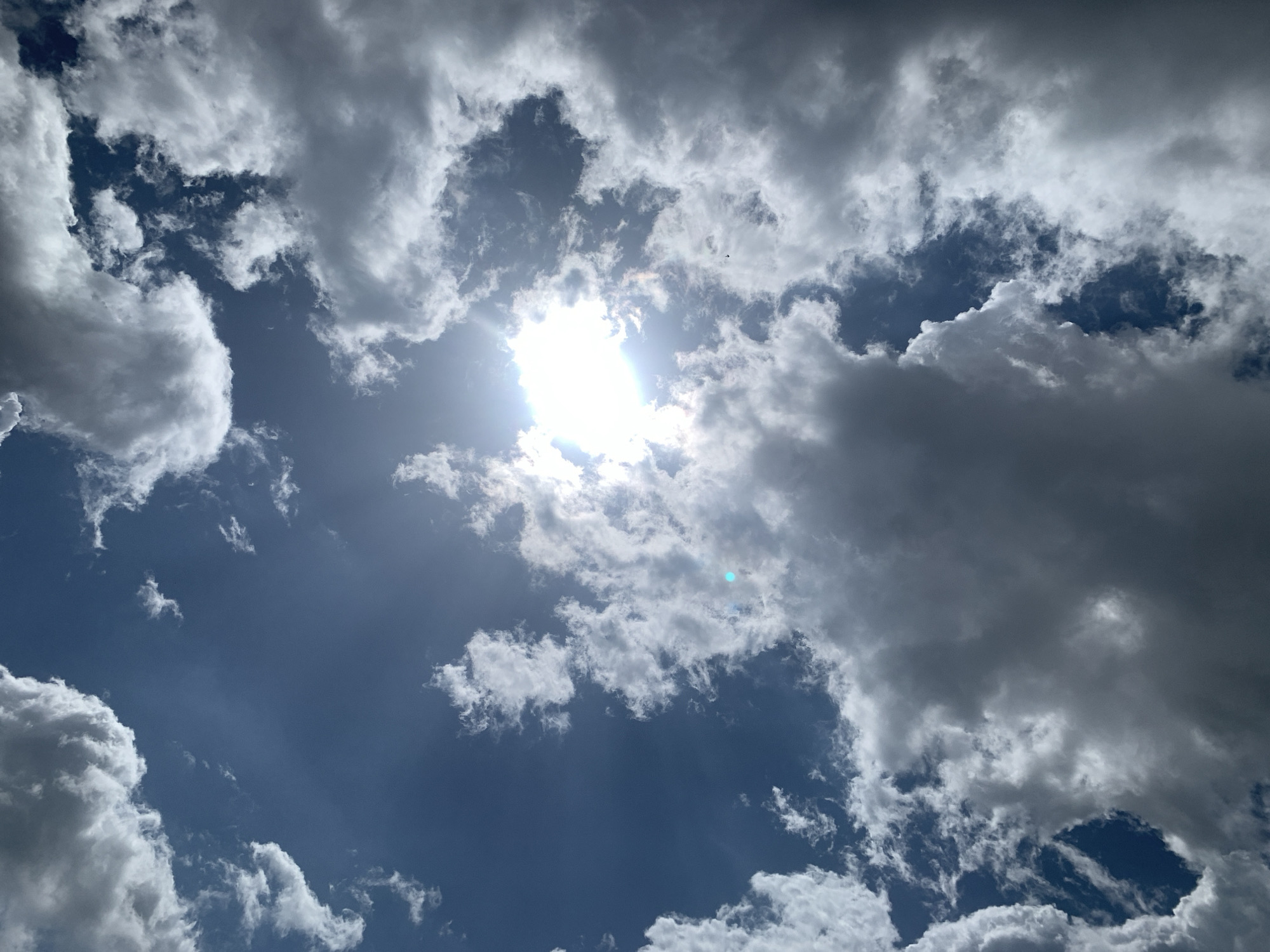 A low-angle, upward-facing shot captures a dynamic sky filled with a mix of dark and light clouds. The sun, a bright and intense point of light, is partially obscured by clouds, casting a brilliant glow and creating sunbeams that pierce through the atmospheric haze. The sky is a deep blue, contrasting with the varying shades of gray and white in the cloud formations. In the distance, a small, dark silhouette of a bird is visible, appearing to fly against the vast expanse of the sky. The overall scene suggests a dramatic and possibly windy day. The image is not specific to Kuru Karama, Nigeria, or any other city.