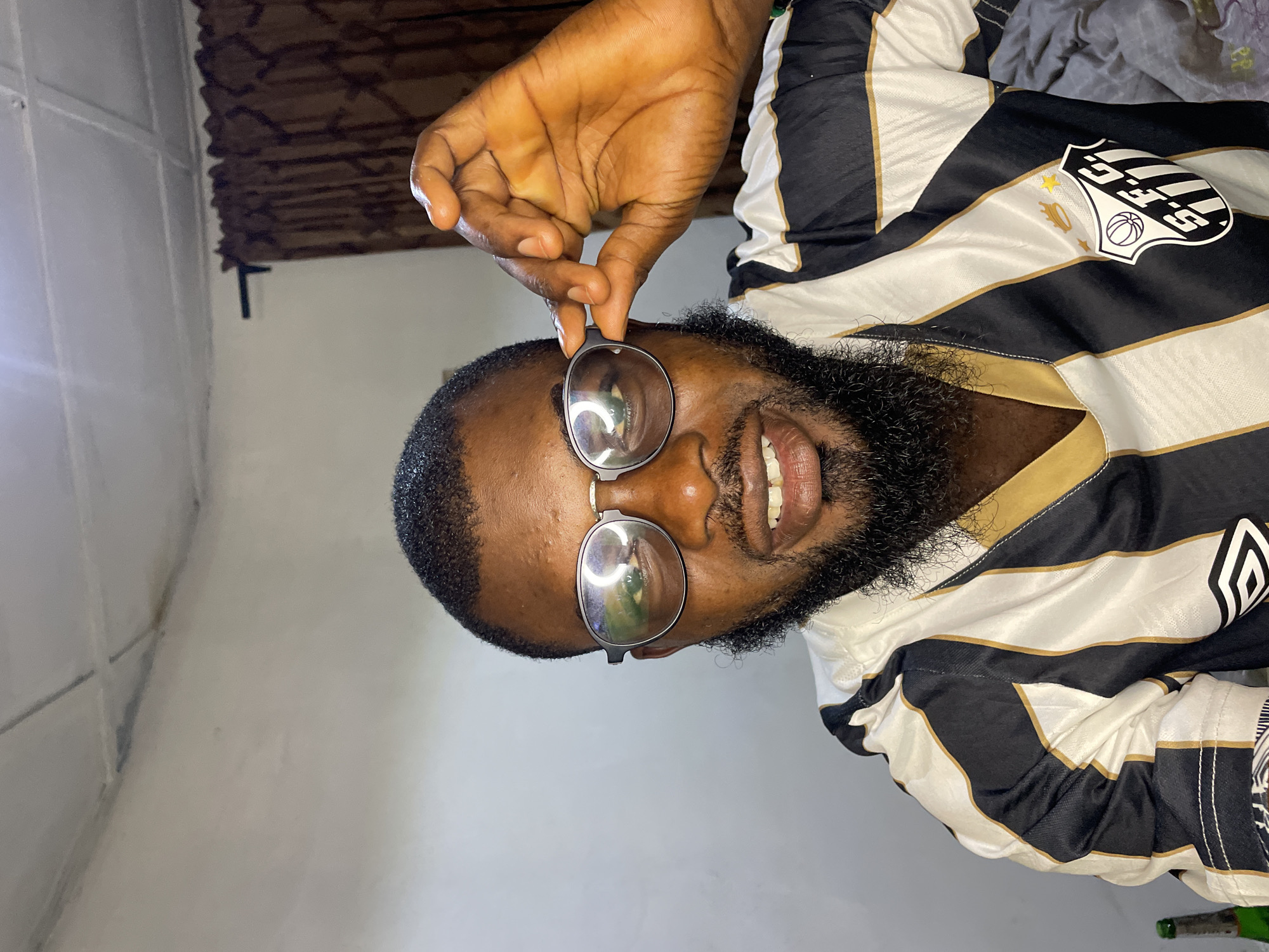 The image shows a close-up, rotated view of a man wearing a black and white striped jersey with gold accents. He has a beard and is wearing round, metallic-rimmed glasses. One of his hands is visible, holding the glasses up to his face. The background suggests an indoor setting with a wall and possibly a window with blinds. The lighting appears to be artificial. There is a crest on the jersey, featuring a shield with a basketball and stars, and the letters "S.F.C." followed by "1912". The overall impression is a casual, personal photograph. The location context is Jos, Nigeria.
