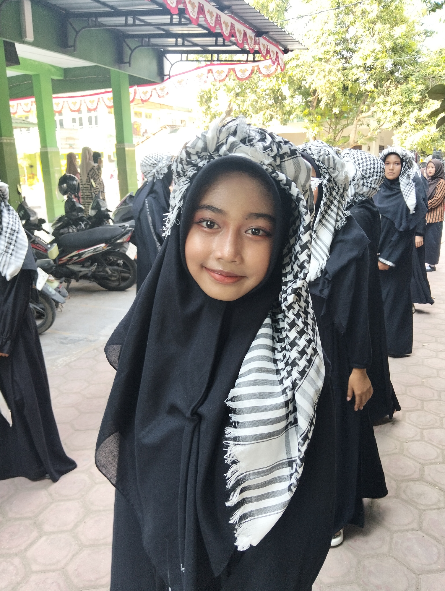 In Kranji, Indonesia, a young woman, the primary subject of the photograph, is positioned in the foreground. She is wearing a black hijab and an elaborate black and white checkered keffiyeh draped over her head, with fringed ends. Her makeup is visible, featuring eyeliner and eyeshadow. She is smiling and looking towards the camera. Behind her, a line of other individuals, also in black hijabs and similar keffiyehs, are visible, some further in the background. Several motorcycles are parked in the mid-ground. The scene appears to be outdoors, with a paved ground and a building with green pillars and a red and white banner in the background. The overall setting suggests a public space or an event in Kranji.