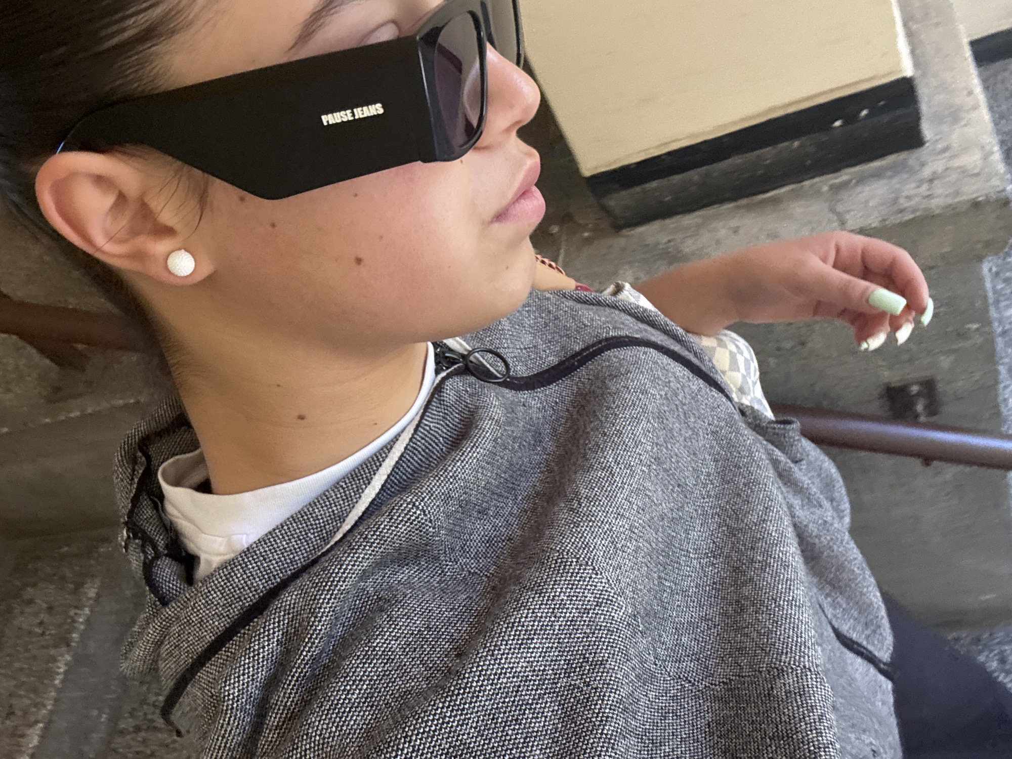 A close-up shot shows a person wearing black sunglasses with the words "PAUSE JEANS" printed on the side. The person has a light complexion and is wearing a gray jacket over a white shirt. They have a round pearl earring in their left ear and manicured nails. The background is a concrete structure, possibly a stairway. The location in the city of Unknown, Unknown is not discernible from the image.