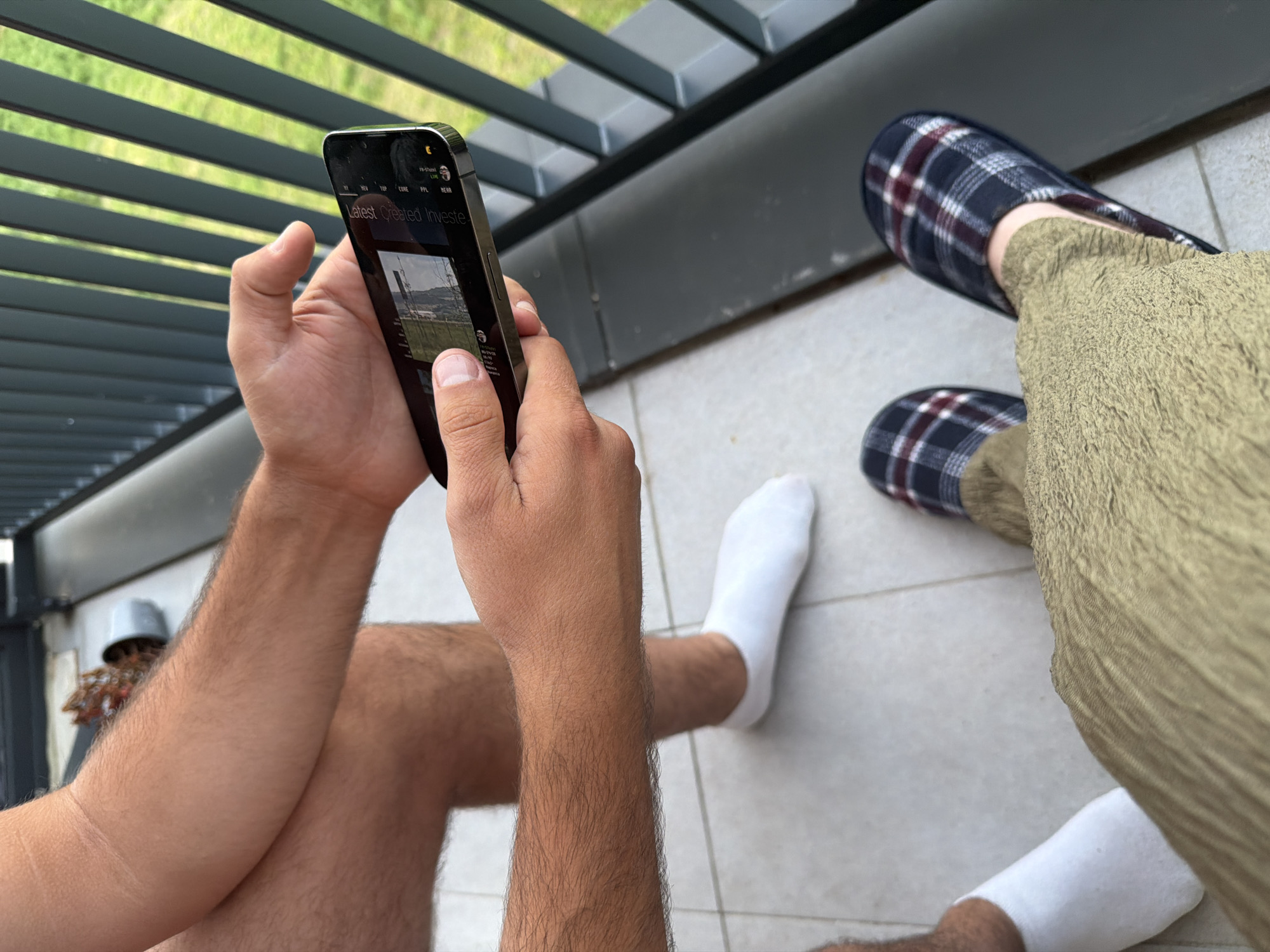 The image shows a close-up perspective of two individuals on a balcony. One person, positioned on the left, is seen from the knees down, wearing white socks. The other person, in the foreground, is seen from the waist down, wearing khaki-colored pants and plaid slippers. This individual is holding a smartphone, actively engaging with it. The phone displays a video or image with text overlay, including "Latest, Created Investe." The background features a railing with vertical gray bars, beyond which is visible green foliage, suggesting an outdoor setting. A potted plant is visible in the background on the left. The flooring of the balcony is made of pale tiles. While the prompt mentions Florești, Romania, there are no specific visual cues within the image to confirm this location.