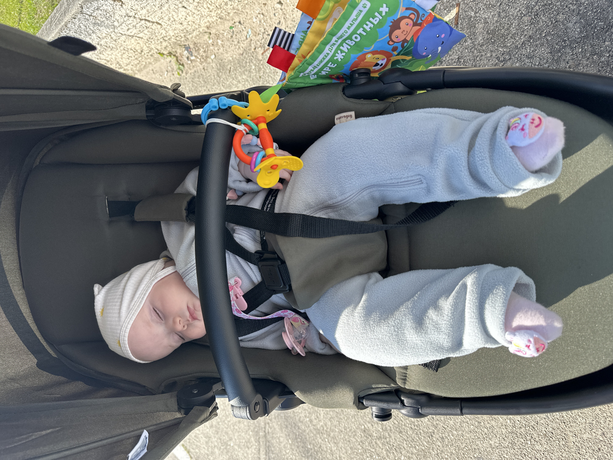 A baby is sleeping in a stroller outdoors on a sunny day. The baby is wearing a white hat with yellow stars and a light blue outfit. A pacifier is attached to the stroller. A toy is also attached to the stroller, and a soft book with animal pictures is hanging from the side of the stroller. The stroller is a dark green color. The background shows a sidewalk and some greenery. The location in Florianópolis, Brazil cannot be determined from the image.