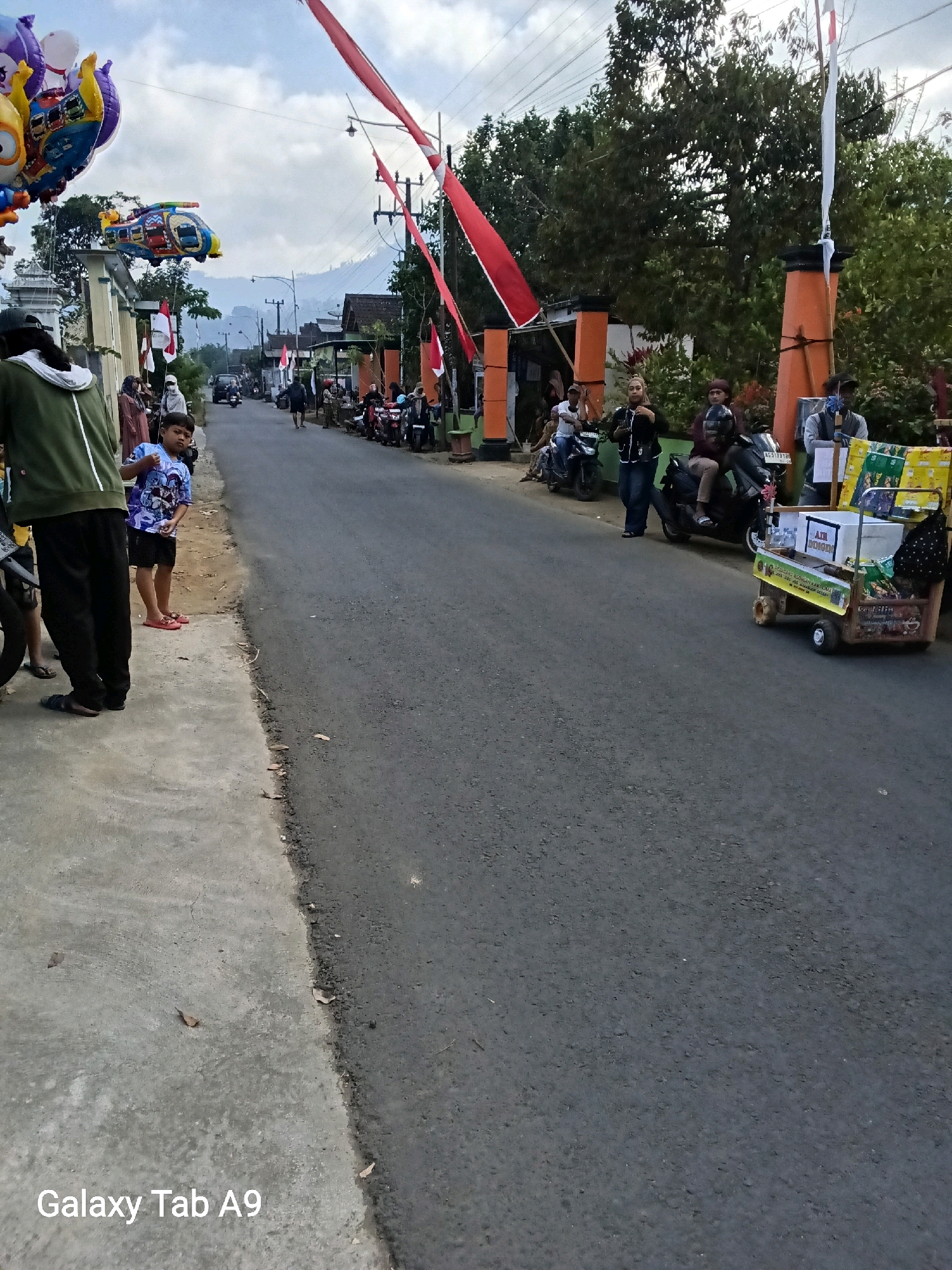 The image depicts a street scene in Bendo, Indonesia, with a focus on a road and sidewalk. Several red and white Indonesian flags are visible, as are colorful balloons shaped like characters and a helicopter. People are standing and sitting along the side of the road, some near parked motorcycles. A small cart with items for sale, including what appears to be bottled drinks, is positioned on the roadside. The sky is partly cloudy, and trees and buildings line the street. The ground surface is asphalt for the road and concrete for the sidewalk.