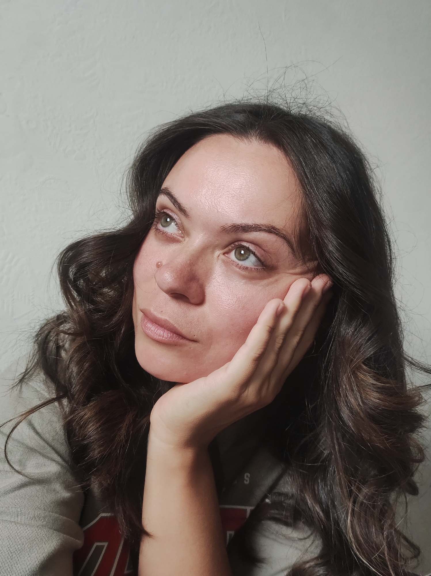 The image shows a close-up, head-and-shoulders portrait of a young woman with long, dark, wavy hair. She is looking upwards and to her left, with her right hand resting on her cheek. Her skin appears to have a light dusting of freckles, and there is a small mole on her right cheekbone. The background is a textured, light-colored wall, typical of interior residential spaces. The woman is wearing a light-colored t-shirt or sweatshirt. There is no indication of the location being in the city of Chaikovsky, Russia, or any other specific geographic location. The image is a still photograph.