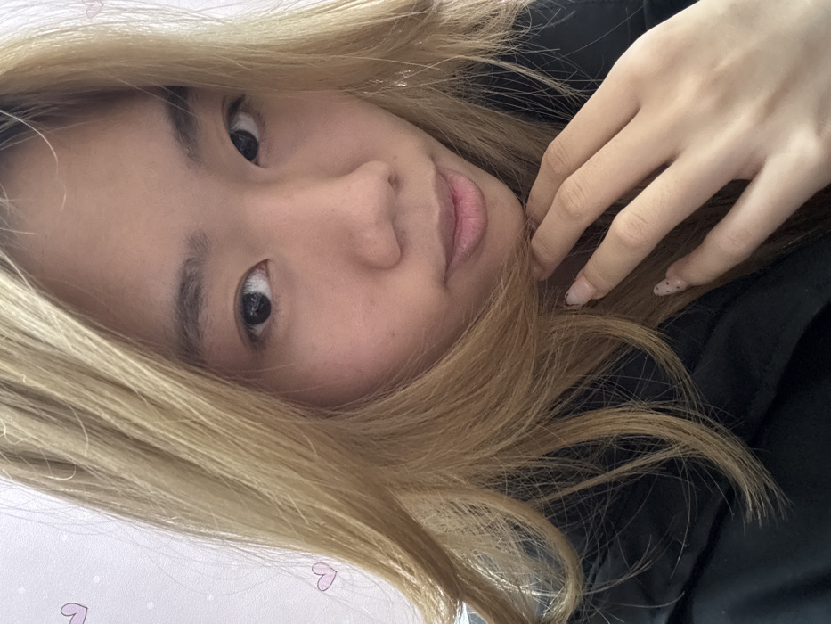 A head-and-shoulder portrait features a young East Asian female looking directly into the camera. She has blonde hair parted on her left side, which cascades over her shoulders and partially obscures her dark, possibly black, attire. Her dark eyes, natural eyebrows, and light skin are visible. Her lips are slightly parted. Her left hand is positioned with fingers slightly curled near her chin and jawline; her nails are manicured with a light pink or nude polish featuring small dark dot patterns. The background is a light-colored, possibly white or pale pink, surface adorned with small, light pink heart graphics and sparse white dots, indicative of an indoor setting. The specific location of Shapingba District, China, cannot be visually confirmed from the image content.