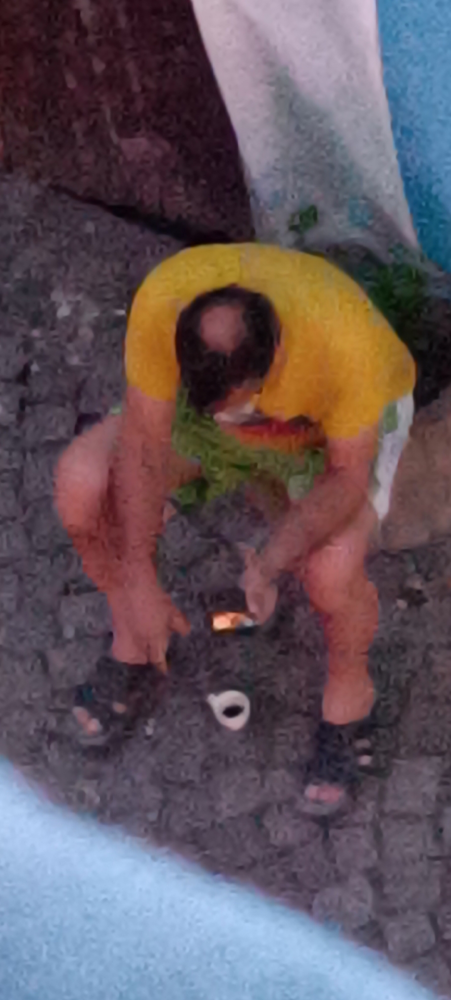 The image, taken from a high angle, shows a bald man with a receding hairline wearing a yellow t-shirt and white shorts. He is crouched down on a cobblestone surface, his legs bent and his back hunched. His right hand is extended, and he appears to be holding a small, rectangular object that is illuminated. His left hand is near the ground, and a white, ring-shaped object is visible in front of his feet. Green vegetation is present behind the man. The scene appears to be outdoors, possibly in a public area, though no specific landmarks of Winnenden, Germany are discernible. The overall image quality is low, with a grainy texture and blurred details.