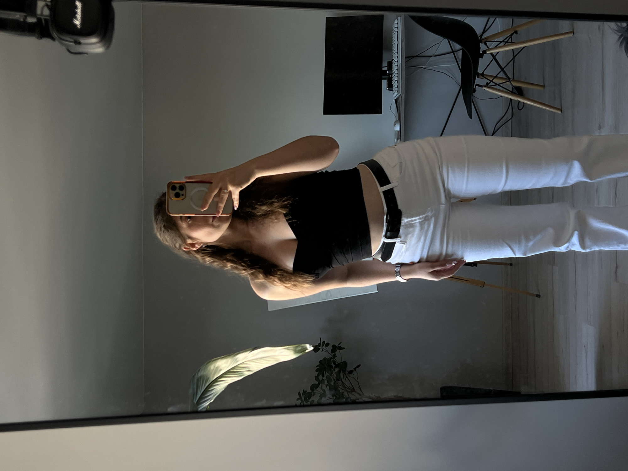 A young woman with long, wavy brown hair is taking a mirror selfie in a room. She is wearing a black, strapless crop top and white high-waisted jeans with a black belt. She is also wearing a watch on her left wrist. In the reflection, a large plant with broad green leaves is visible to her left. To her right, a desk with a computer monitor, keyboard, and mouse is visible, along with a black chair with wooden legs. Above the desk, a pair of black headphones with "Marshall" written on them are hanging from something unseen. The background walls are a neutral gray. The flooring appears to be light-colored wood. The overall environment suggests an indoor domestic setting, possibly a bedroom or living area. There are no specific landmarks or clues that would indicate the location being Ternopil, Ukraine.