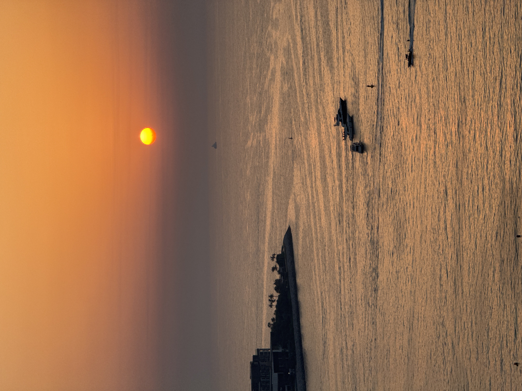 This is a high-angle, vertical shot of a sunset over the Arabian Gulf in Dubai, United Arab Emirates. The sun is a bright yellow orb near the horizon, casting a warm orange glow across the sky and water. Several yachts are scattered across the water, leaving white wakes. A cluster of buildings and palm trees are visible on a man-made peninsula in the foreground, likely part of a residential or resort development. The water is calm, with gentle ripples reflecting the sunset's colors. A distant cargo ship can be seen on the horizon. The scene evokes a sense of tranquility and luxury associated with Dubai's coastal lifestyle.