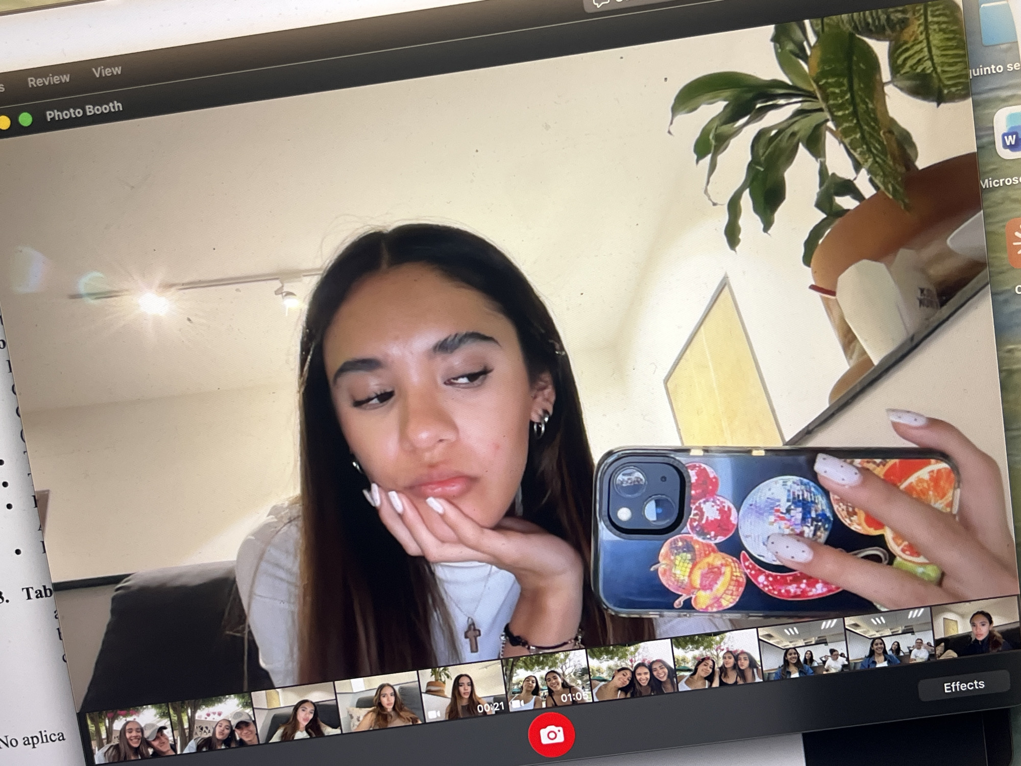 The image shows a screenshot of a computer screen. At the top of the screen is the menu bar of an Apple computer with the words "Review" and "View" visible, and an active window labeled "Photo Booth". The main part of the screen shows a young woman with long, dark hair, fair skin, wearing a white shirt and jewelry, and posing with her hand under her chin in a possible photo booth preview. She is holding a cell phone in front of the camera lens, and it has a clear case decorated with fruits and a disco ball image. In the background of the image is a room with neutral colored walls, a framed piece of art, and a potted plant. At the bottom of the screen are thumbnails of images of people and a red record button. To the left of the screen, part of a white document is visible with text in Spanish.