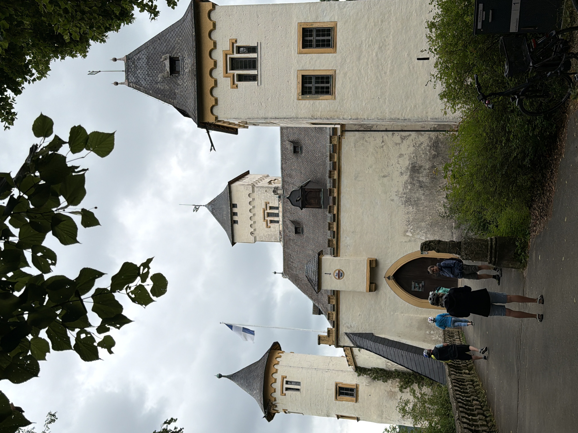 The image shows a cloudy day at the Heiligenstein Castle in Heiligenstadt in. OFr., Germany. A group of people, some wearing helmets, are walking on a path leading to the castle's entrance. The castle itself is a cream-colored building with several towers, a prominent flag flying from a flagpole, and dark-tiled roofs. The foreground includes green foliage and a parked bicycle.