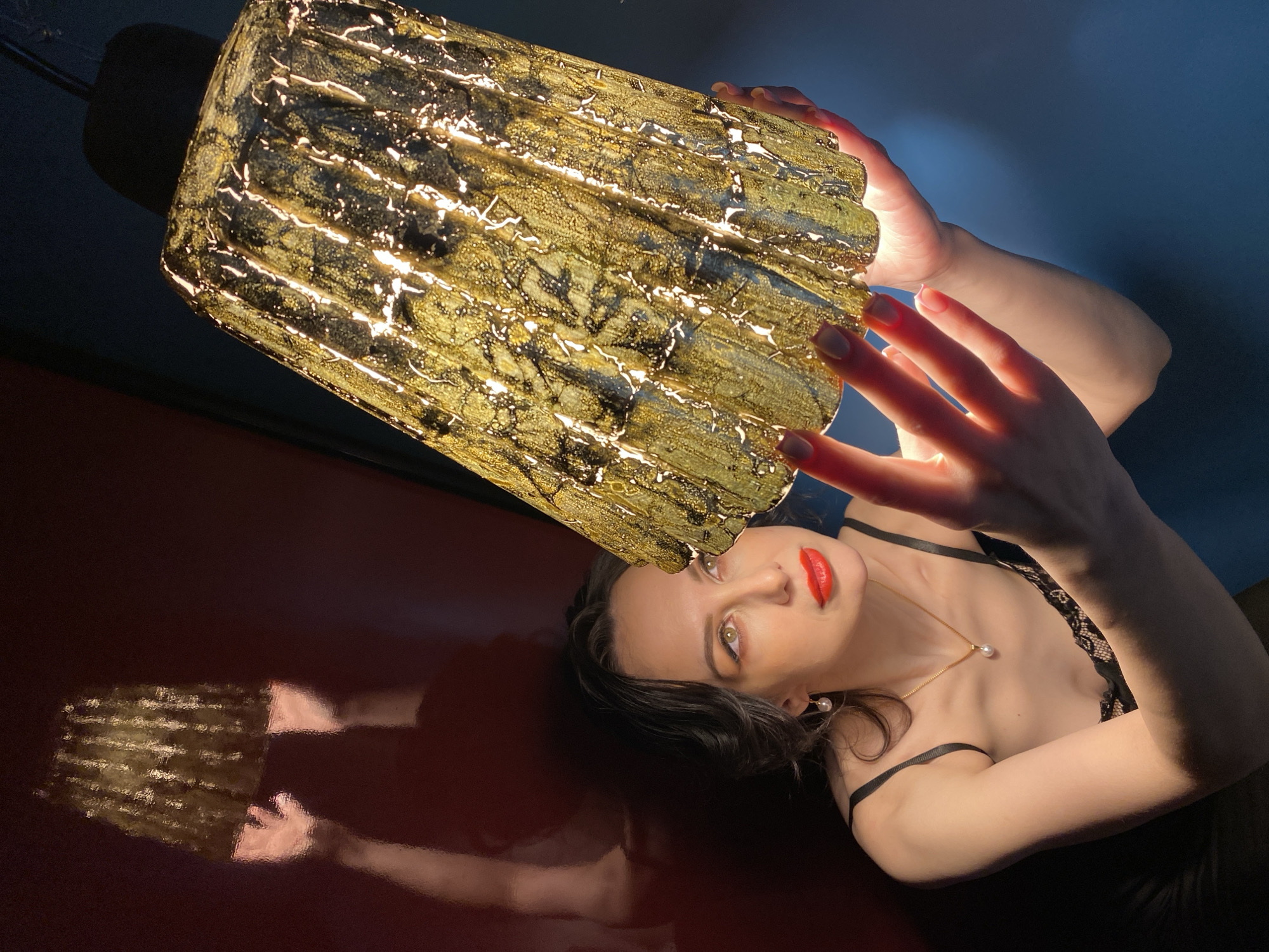A woman with fair skin, red lipstick, and dark hair lies on a shiny, dark red surface under a lit, cylindrical, textured gold lamp. She is wearing a black top with thin straps and a gold necklace with a pearl pendant. Her left hand is raised towards the lamp, fingers spread. The background transitions from red to blue. The reflection of the lamp and hands are visible on the red surface. It is unlikely to ascertain this was taken in Yekaterinburg, Russia, without additional information.