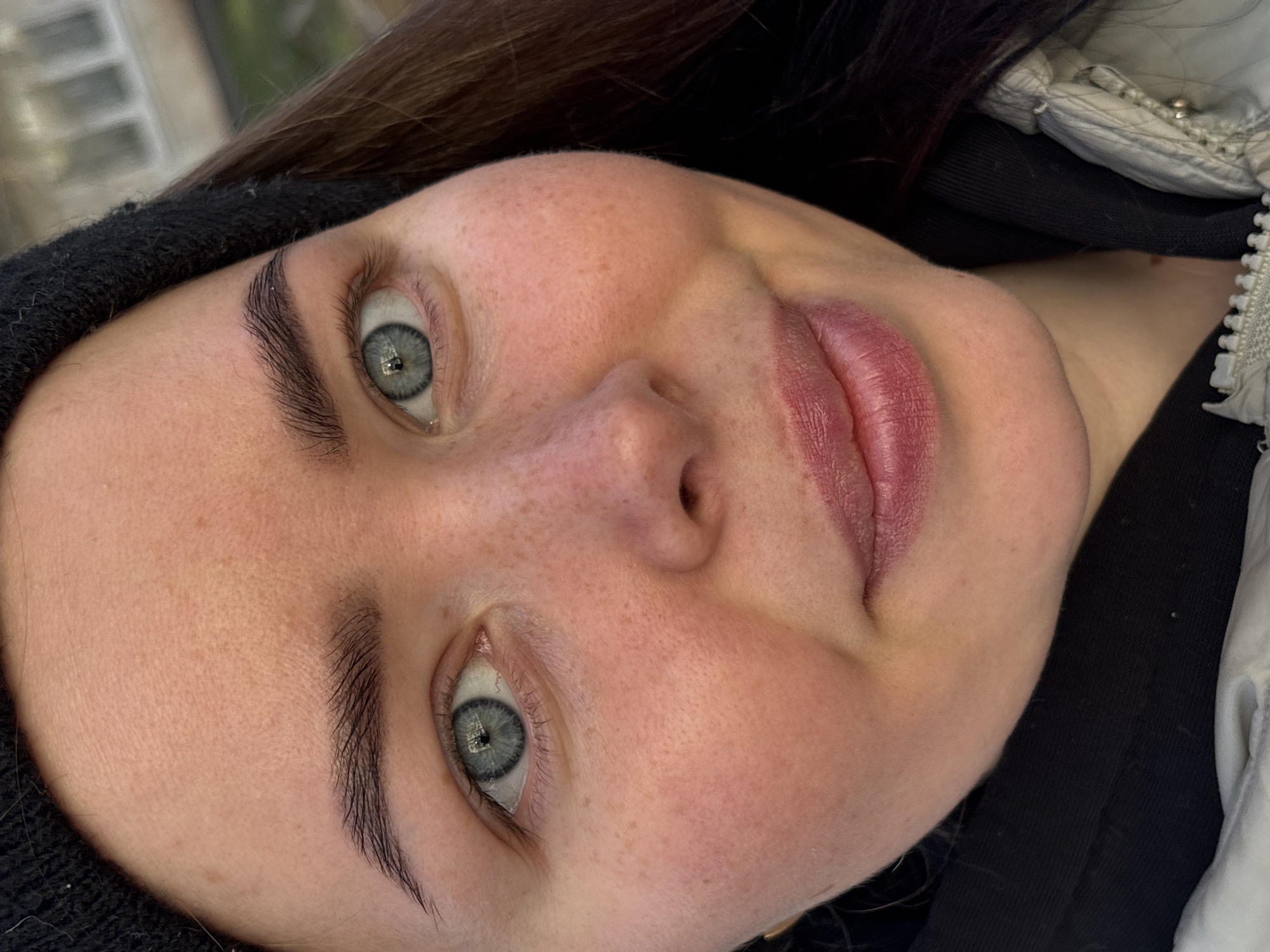 A close-up, high-angle portrait shot features a young individual, likely female, with the head slightly tilted and turned, looking directly into the camera. The subject has fair skin with numerous light freckles visible across the nose and cheeks. Her prominent eyes are a striking light blue-grey color with dark pupils and intricate iris patterns. Her eyebrows are dark, well-defined, and appear meticulously shaped or styled, with individual hair strokes clearly visible. The lips are full, a natural rosy pink, and show fine vertical lines suggesting natural texture. Dark hair frames the top of her face, partially covered by a black knit hat or beanie. She is wearing a dark long-sleeved garment under a light-colored (possibly white or light grey) puffer jacket, the zipper and collar of which are visible on the right side of the frame. The background is softly blurred, showing indistinct shapes, suggesting natural lighting, possibly an outdoor or brightly lit indoor setting. The expression is neutral. This image is associated with Izhevsk, Russia.