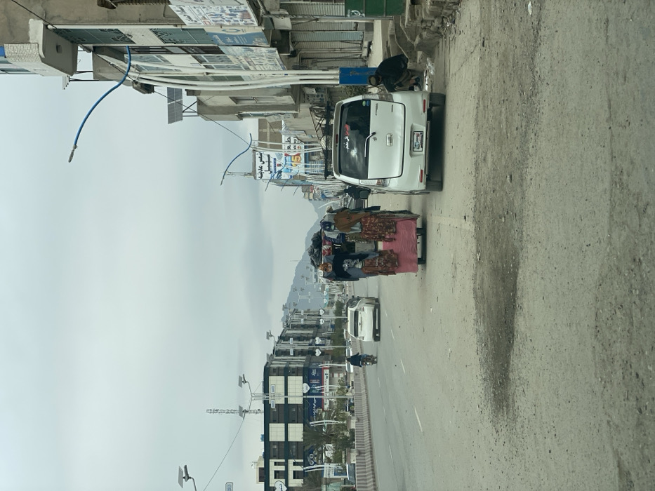 This image captures a street scene in Kandahar, Afghanistan, under an overcast sky. The foreground depicts a dusty, somewhat uneven asphalt road with faint white lane markings.
In the center of the image, a two-wheeled pushcart, approximately chest-height, is positioned in the right lane. It features a red/pink base and is laden with various goods. Bundled textiles and articles of clothing, resembling dresses or abayas, are visibly hung from frames on the cart, while other items are piled on top, partially covered by a dark tarp.
To the immediate right of the pushcart, a white compact hatchback vehicle, possibly a Suzuki Wagon R, is parked or stationary in the same lane. It has a visible license plate and some Arabic script on its rear window. Further right, along the dirt shoulder and curb, a male individual dressed in dark traditional attire (likely shalwar kameez) is seated on a low concrete step or raised curb, facing partially left. Green waste bins are visible further to the right on the curb.
The right side of the street is lined with multi-story buildings, some featuring commercial storefronts, many with closed metal shutters. Numerous vertical white conduit pipes are visible on the building exteriors. Signs with Arabic/Dari script are affixed to the buildings and on large banners, including one reading "سنس مدينة" (city sense/center). Solar-powered streetlights with curved blue elements extend over the road from poles along the sidewalk.
On the left side of the street, a multi-lane thoroughfare is separated by a low concrete median barrier. A person on a motorcycle is visible in the far left lane, moving away from the viewer, followed by a white sedan. Beyond the median, more multi-story buildings with modern-style dark and light paneling line the street. Additional solar-powered streetlights are present along the median. In the distant background, under the grey sky, hills or mountains are discernible.