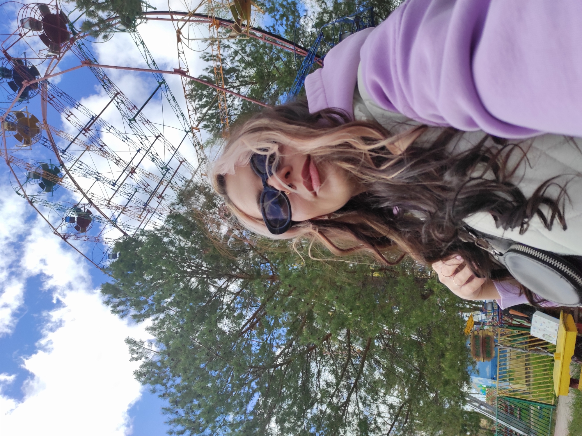 The image is a selfie taken at an amusement park in the city of Chaikovsky, Russia. A woman with blonde-streaked brown hair, wearing sunglasses and a light purple hooded sweatshirt, is the main subject. Behind her, a Ferris wheel stands against a backdrop of blue sky with scattered clouds. Trees are visible, partially obscuring the amusement park attractions in the background. A portion of a yellow railing and what appears to be a section of a painted wall or structure are visible on the right side of the frame. The woman appears to be posing for the photograph.