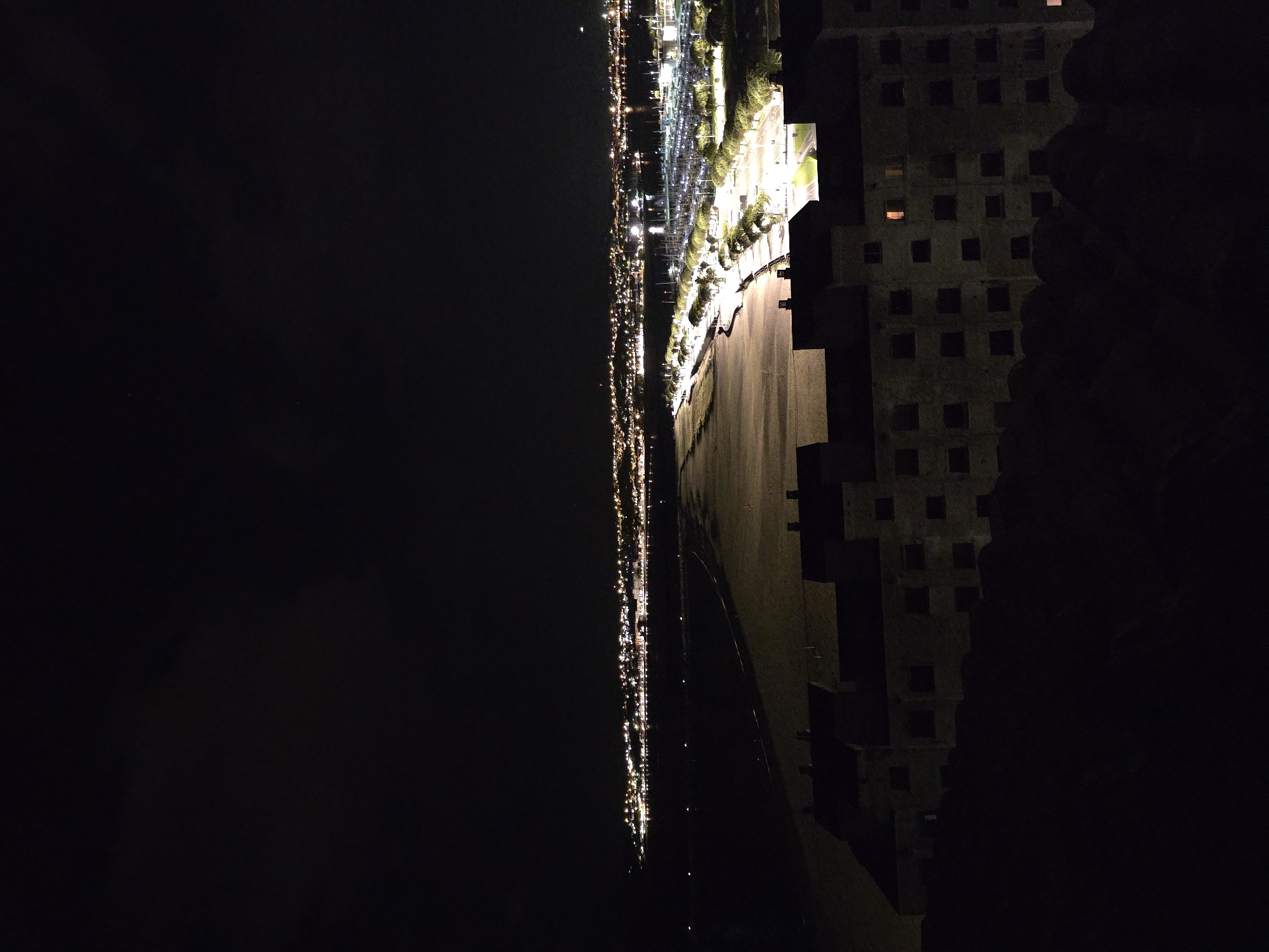 The image shows a night view of Hondarribia, Spain, from a high vantage point. To the right is a building with multiple windows. The foreground appears dark, possibly trees and rocks. The lit-up area of the city is on the left. There is a large illuminated section of a parking area or port, with rows of lights and possibly vehicles. Beyond that, a line of lights indicates a waterfront or road along the water's edge, stretching far into the distance. The rest of the scene is shrouded in darkness.