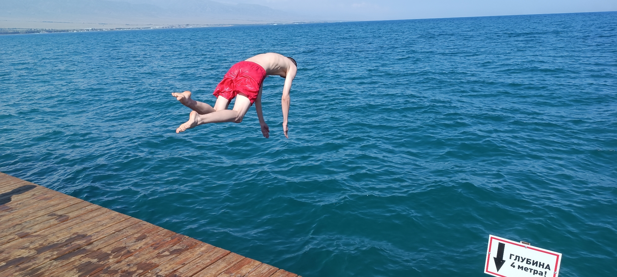 A young man in red swim trunks is captured mid-air, in the process of jumping into a body of water. His body is angled downwards, with his arms extended and legs bent. He appears to be jumping from a wooden pier that occupies the left side of the frame. The water is a vibrant blue, with small ripples on its surface. In the background, a distant shoreline is visible with some vegetation and buildings, all under a clear blue sky. A white sign with red trim and black text pointing downwards is partially submerged in the water on the right side of the image.