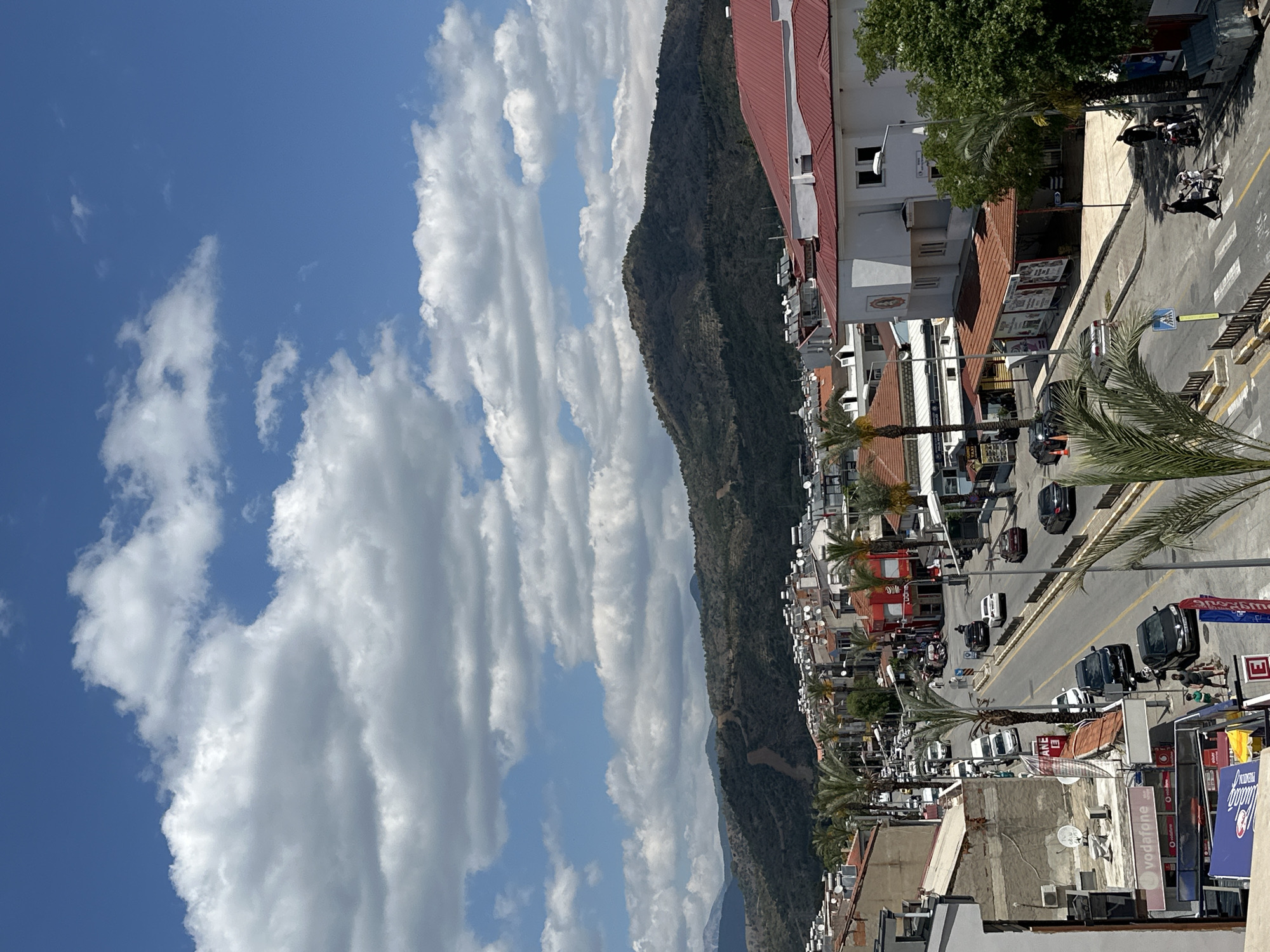 The image presents a high-angle view of a street scene in Fethiye, Turkey. A mountain rises in the background, stretching behind a line of buildings. Cumulus clouds fill a predominantly blue sky. A street runs through the city, with palm trees lining the sidewalks. Cars move along the road, and pedestrians cross at designated crosswalks. Buildings of various heights and architectural styles populate the street edges, with some featuring red-tiled roofs. Signage for Vodafone and other businesses is visible, indicating a commercial district. Satellite dishes are mounted on the roofs of several buildings.