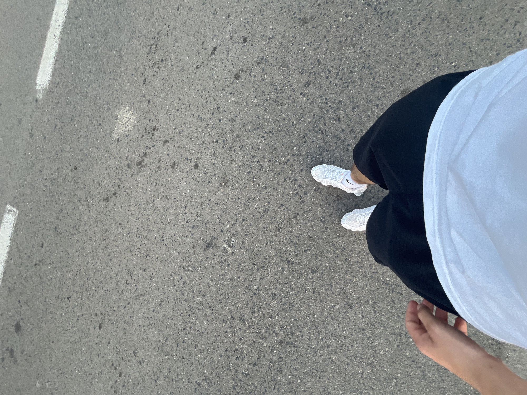The image shows a first-person perspective of someone walking on asphalt pavement. The person is wearing black shorts, a white t-shirt, and white sneakers. A hand is visible on the left side of the frame. There are two white painted lines on the asphalt, indicating a road marking. The scene appears to be outdoors, possibly in a street or pathway. The location is identified as Vladimirovca, Moldova.