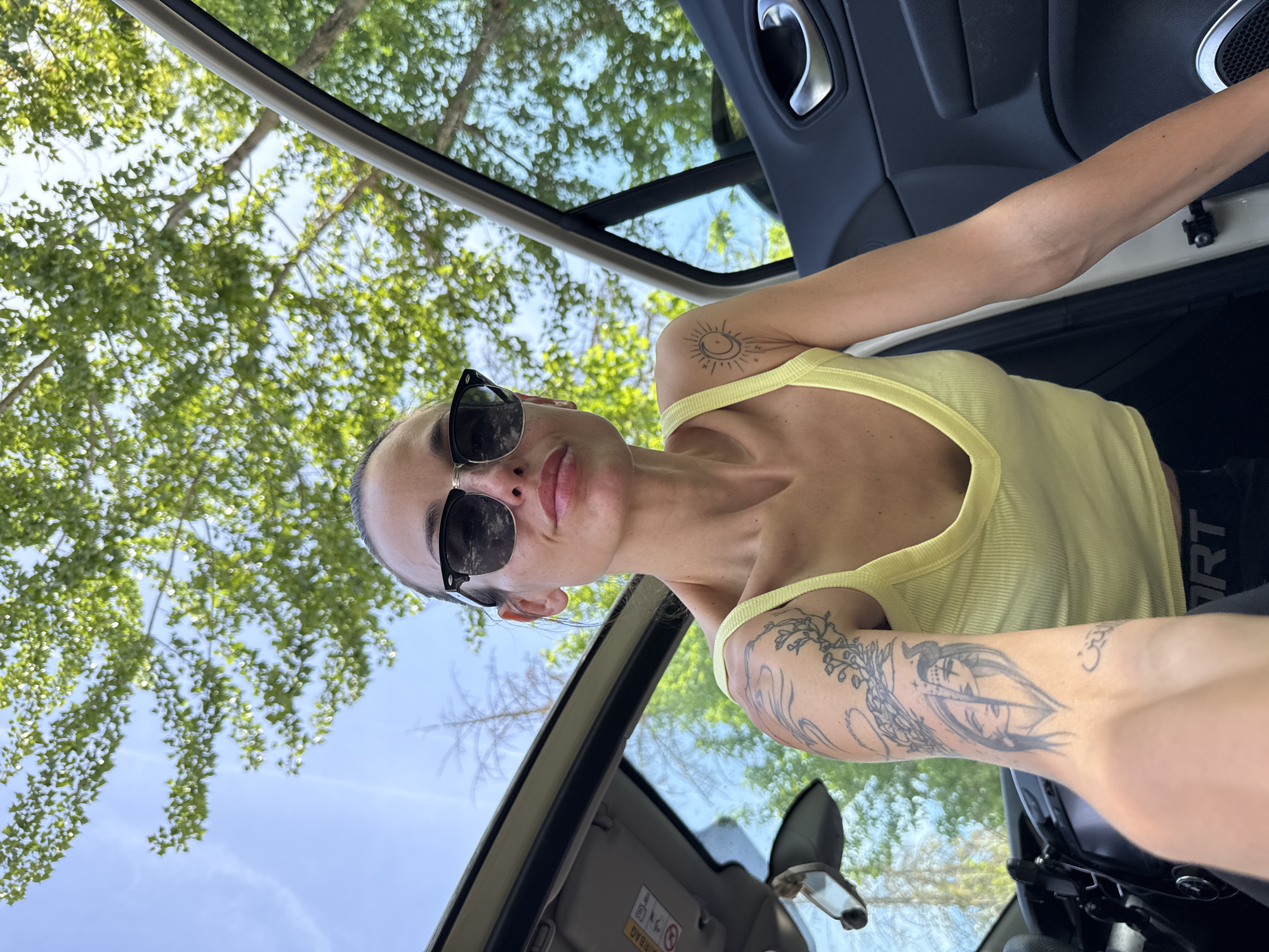 The image displays a rotated, selfie-style portrait of a person inside a vehicle, looking upwards towards the camera. The subject is a light-skinned individual wearing black sunglasses and a yellow ribbed tank top. Their head is tilted back, with a neutral expression and slightly parted lips. Visible tattoos adorn both arms: a stylized sun and moon design is on the upper right arm, while the left forearm features intricate floral and leafy patterns, along with what appears to be a face and a small symbol resembling "Om."
The setting is the interior of a car, with dark door panels, a metallic door handle, and a sun visor visible. The word "AIRBAG" is clearly printed on the sun visor. On a piece of dark clothing partially visible on the subject's right side, the word "SMART" is present. Through what appears to be an open sunroof or convertible top, a bright blue sky with some light clouds is visible, along with the vibrant green canopy of trees, indicating a clear, sunny daytime. The scene captures a relaxed moment, likely in Pleven, Bulgaria.